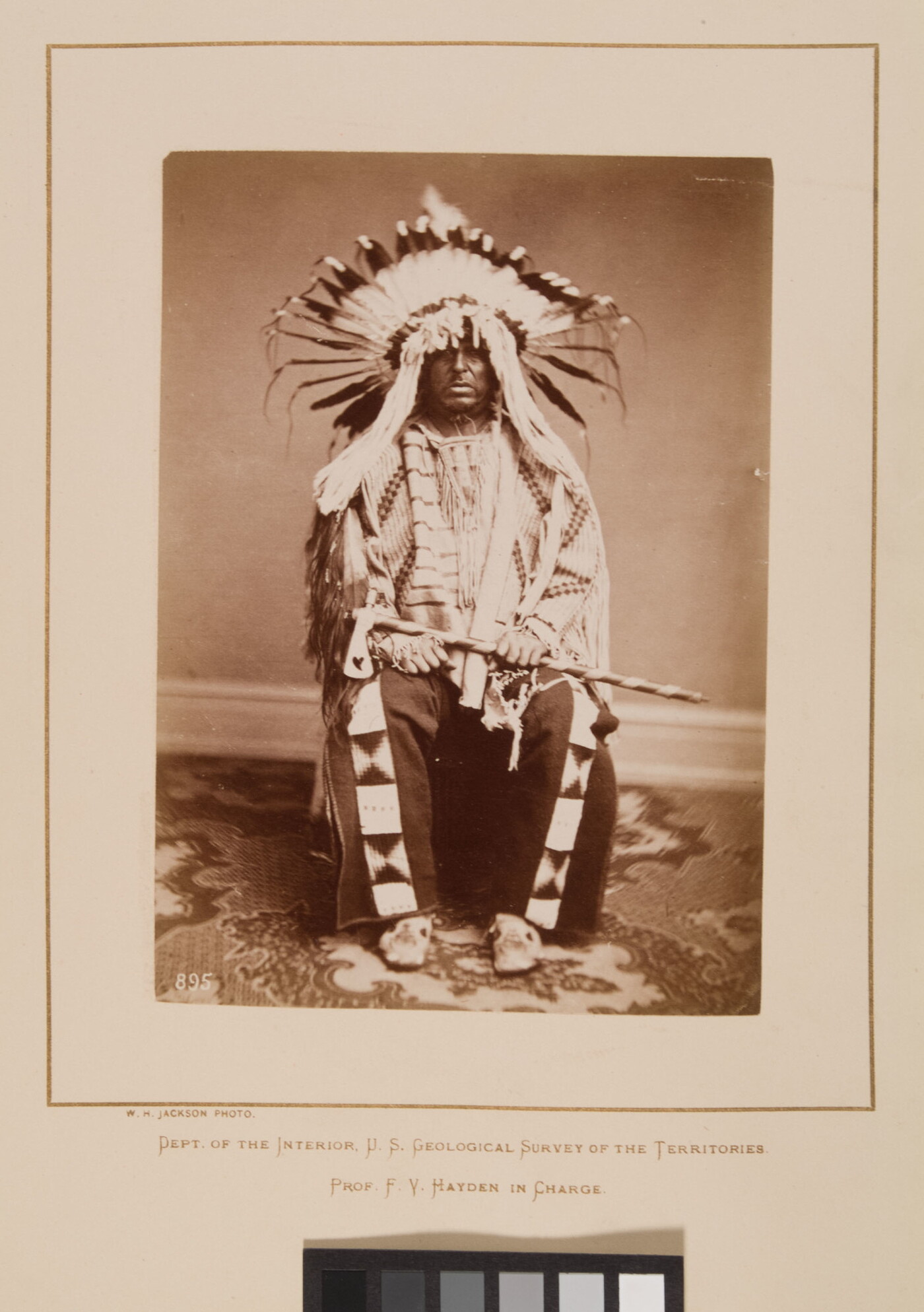 A black-and-white formal portrait photograph of a seated Indigenous man in full, traditional regalia with a headdress and tomahawk.