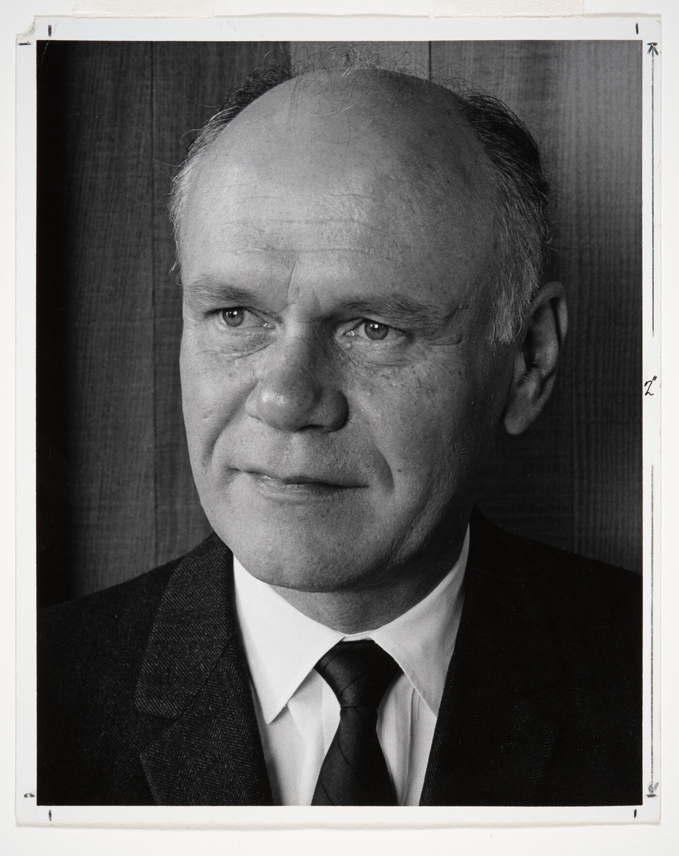 A black-and-white portrait photograph of a mostly bald White man gazing to the side wearing a dark jacket, dark tie, and white shirt.
