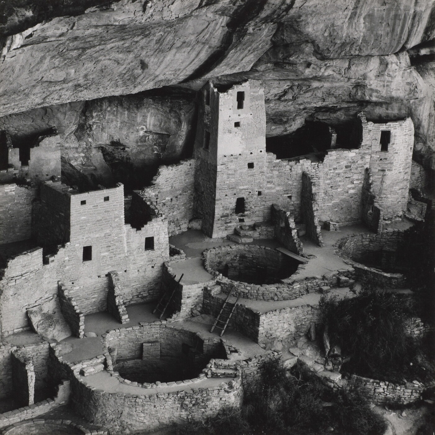 A black-and-white photograph of ruined stone buildings, some with towers, and several underground circular rooms all under the overhang of a cliff.