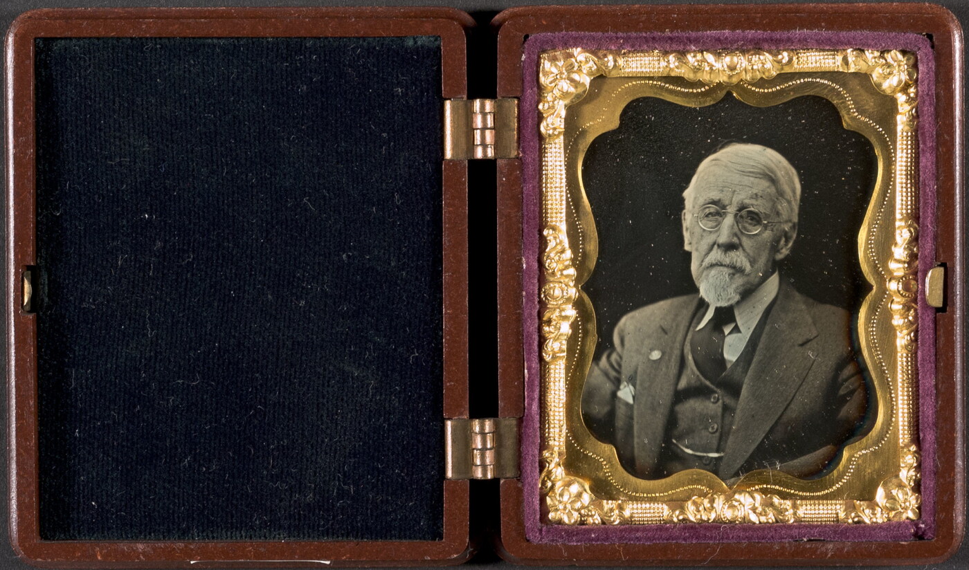 A black-and-white portrait photograph in a case with a decorative gold frame of an older, White man in a suit and glasses.