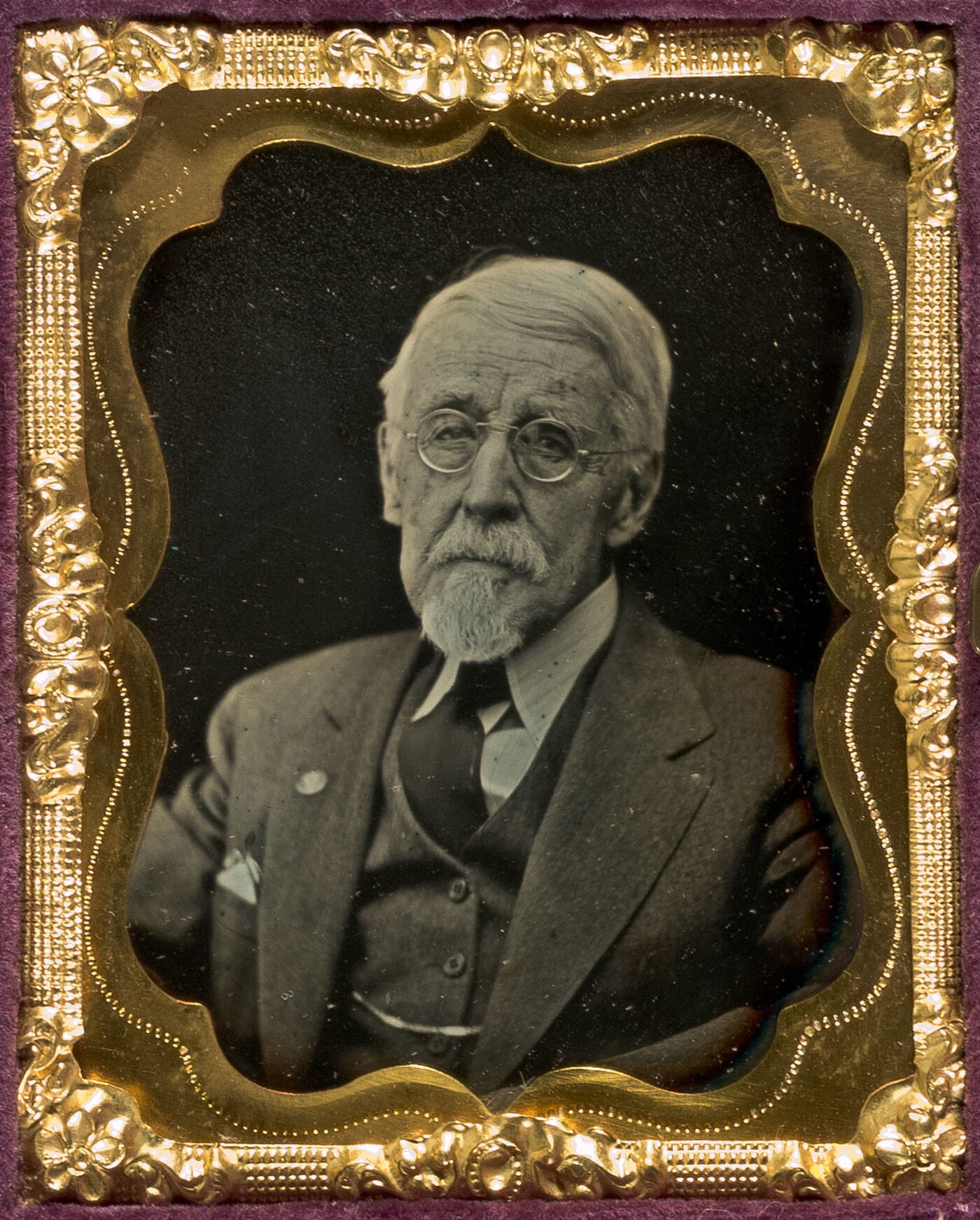 A black-and-white portrait photograph in a decorative gold frame of an older, White man in a suit and glasses.