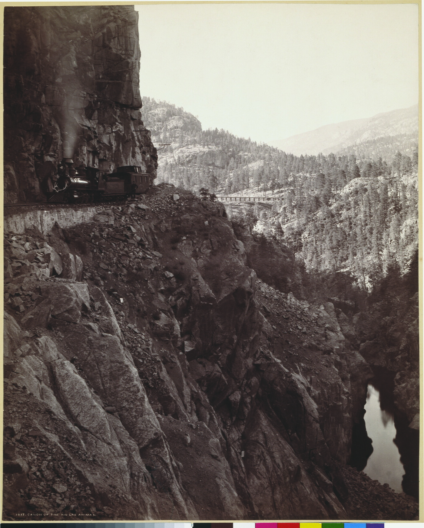 A sepia-toned photograph of a train travelling on the edge of a rocky, mountainside cliff.