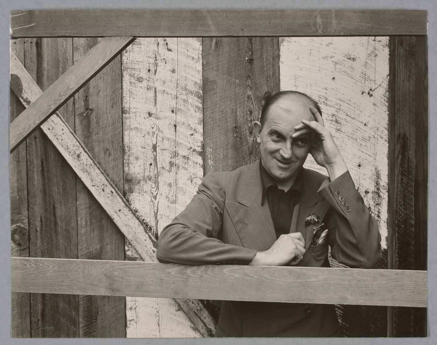 A black-and-white photograph of a bald, White man standing next to a wood structure, wearing a suit jacket and resting his head in his hand.