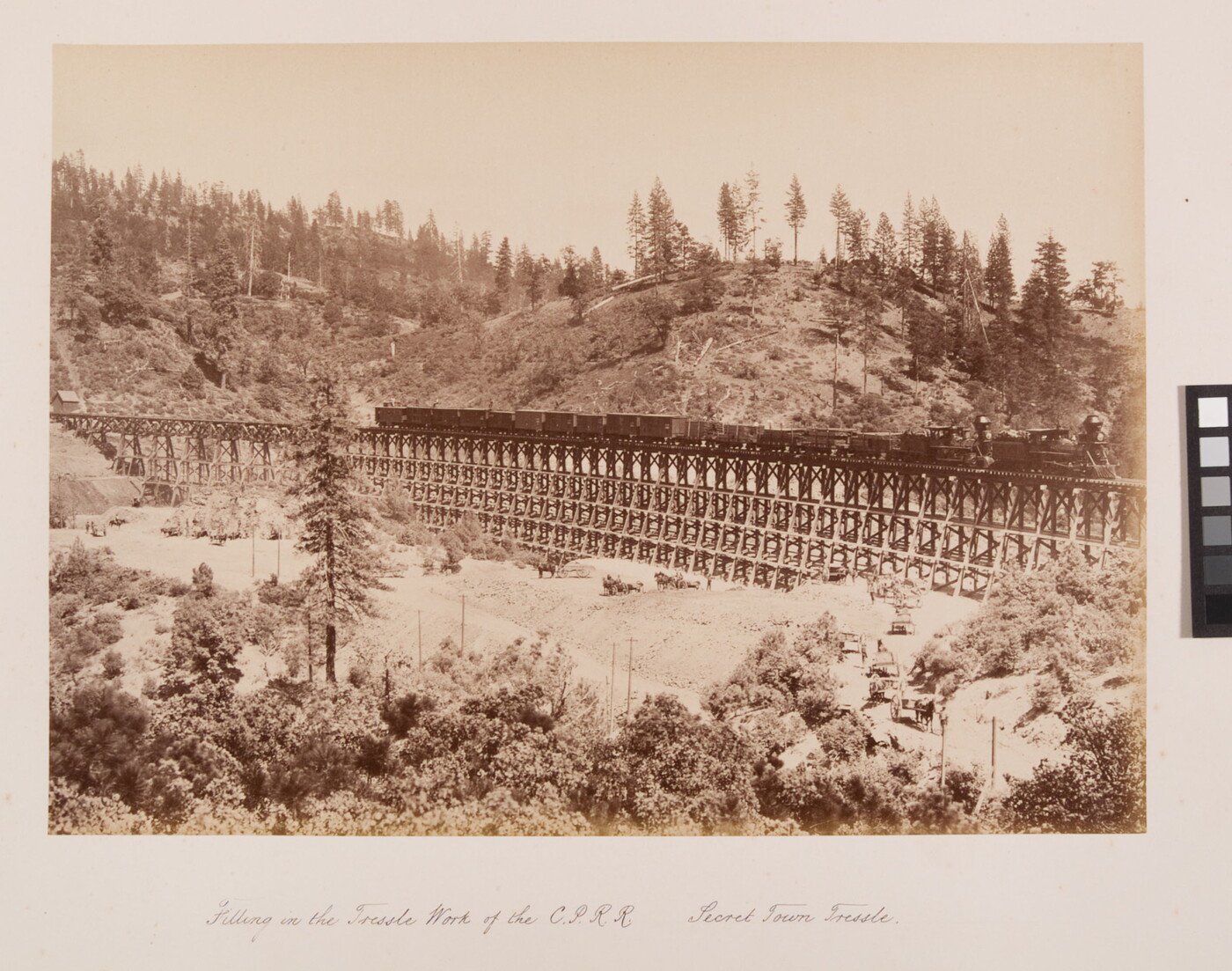 A sepia-toned photograph of a train travelling across a mountain valley with onlookers at base of the trestle.