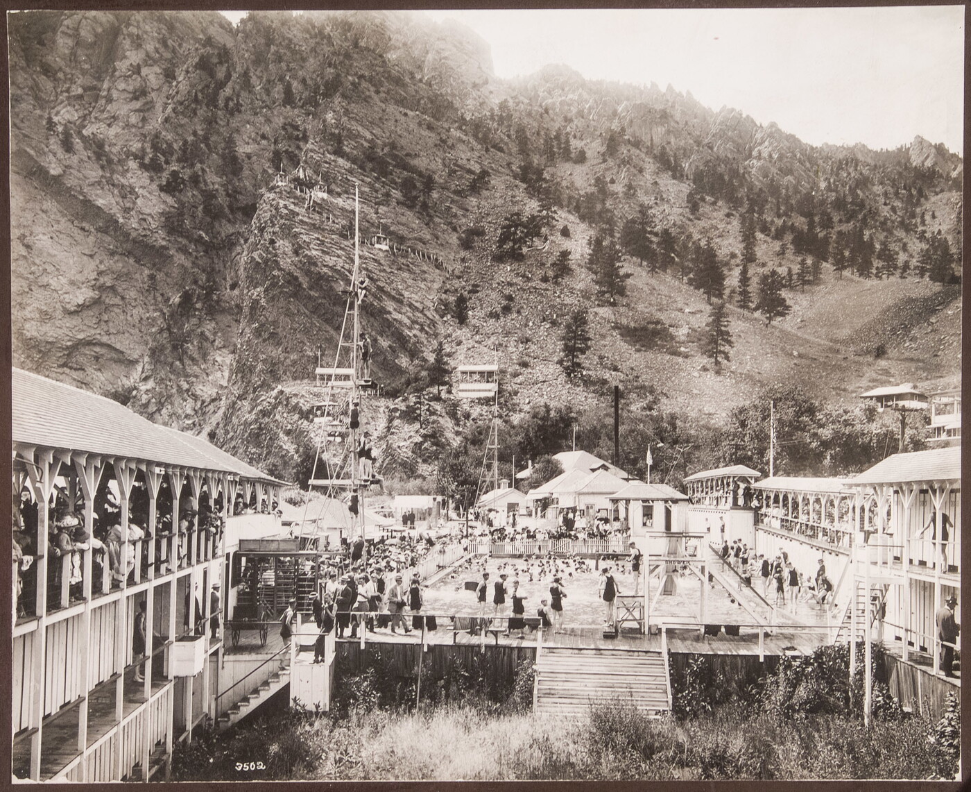 A black-and-white photograph of many people swimming in a large pool nestled at the base of a mountain.