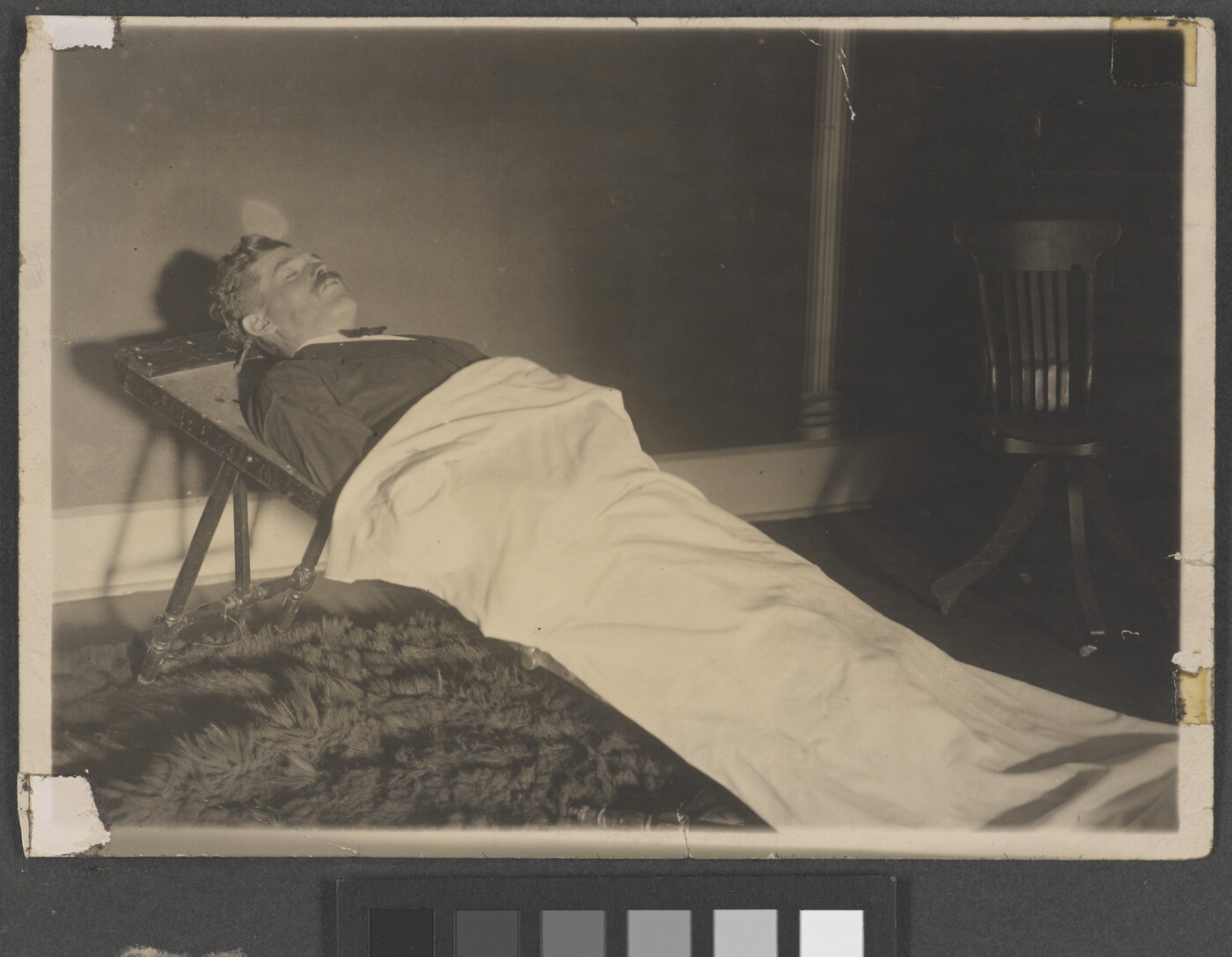 A black-and-white photograph of a White man in a formal suit laying on a stretcher and partially covered by a white sheet.