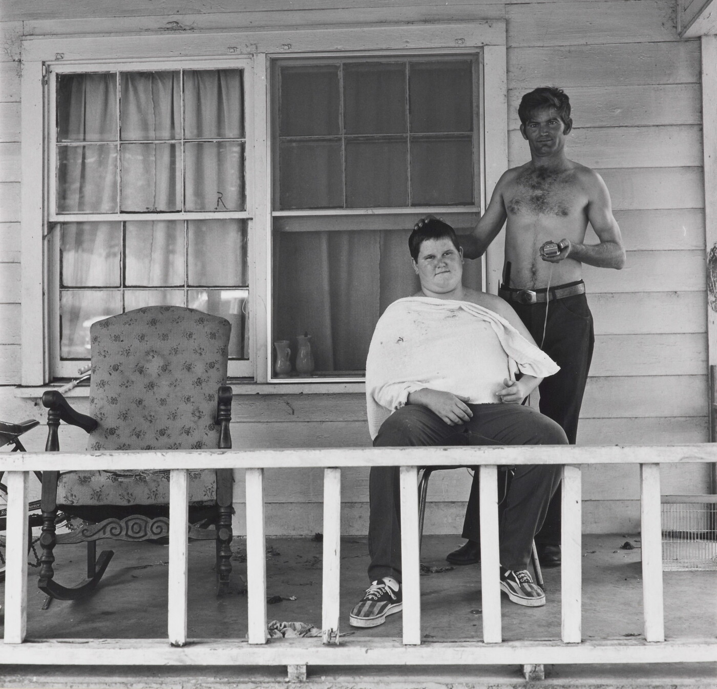 A black-and-white photograph of two White men on the porch of a house: one seated with a white apron around his neck and shoulders; the other standing, shirtless, holding clippers in one hand, the other on the seated man&#039;s head.