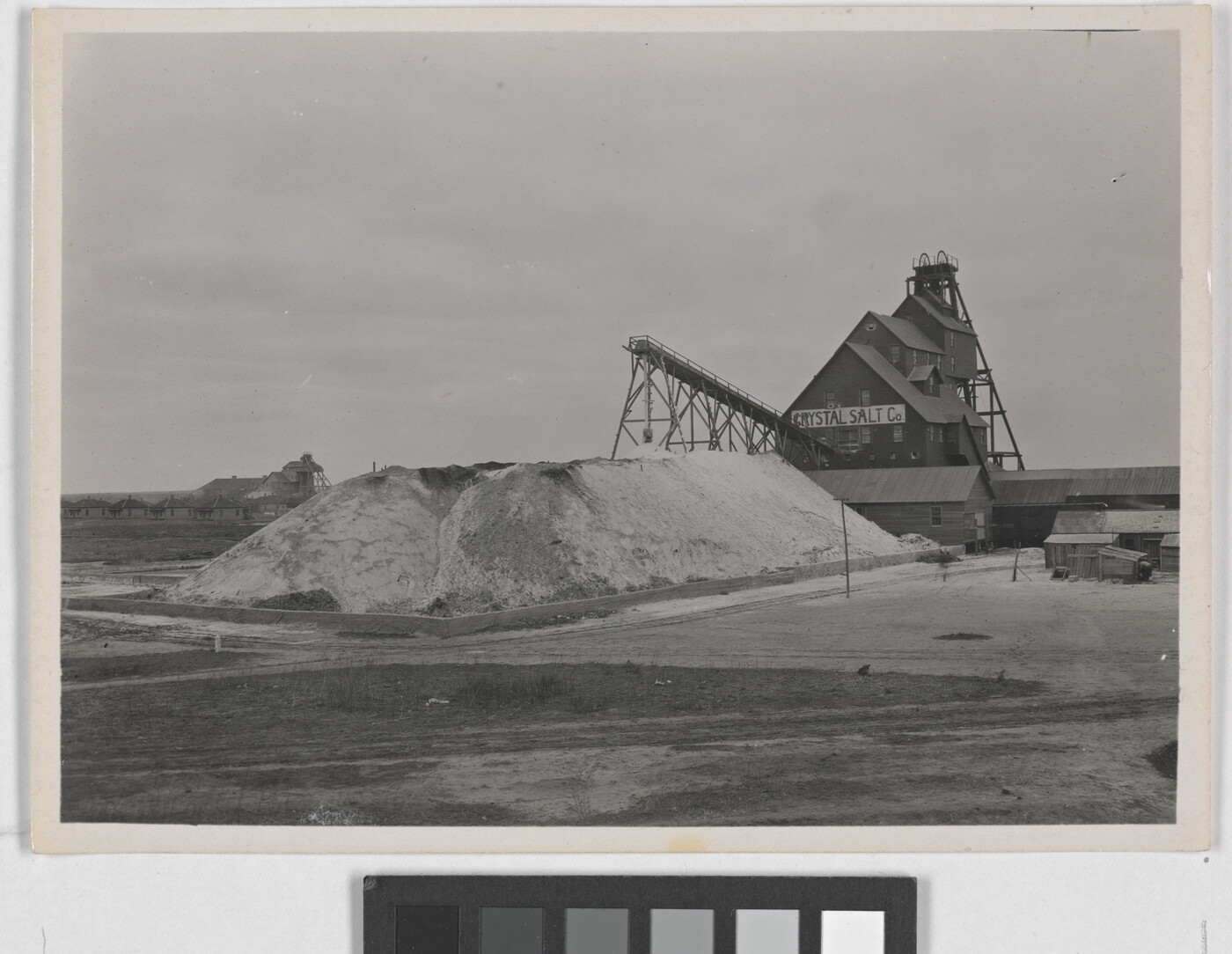 A black-and-white photograph of large pile of salt in front of a multi-story industrial building with a sign that reads &quot;Crystal Salt Co.&quot;