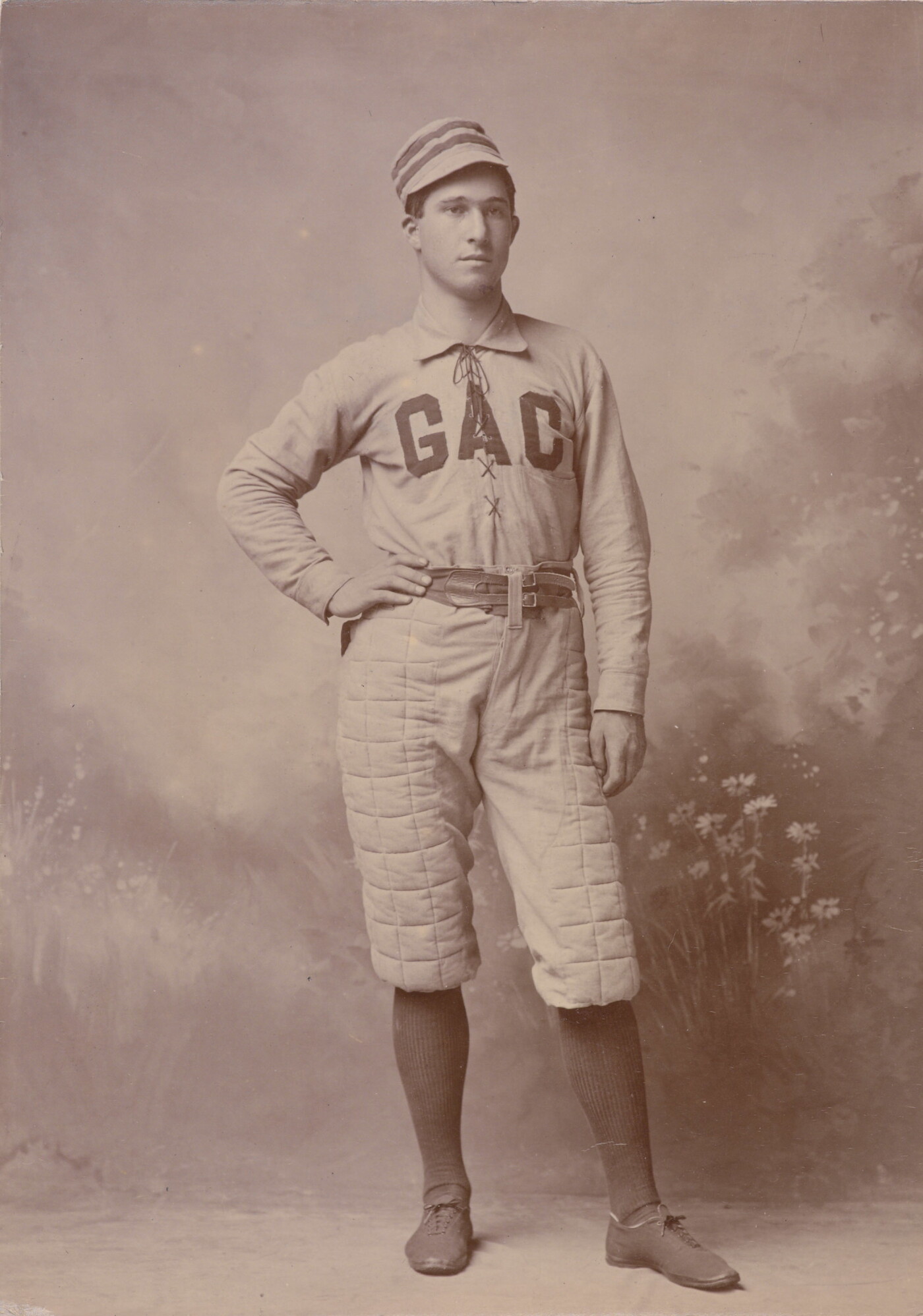A sepia-toned portrait photograph of a White man, one hand on hip, wearing a baseball hat and uniform with &quot;GAC&quot; across the chest.