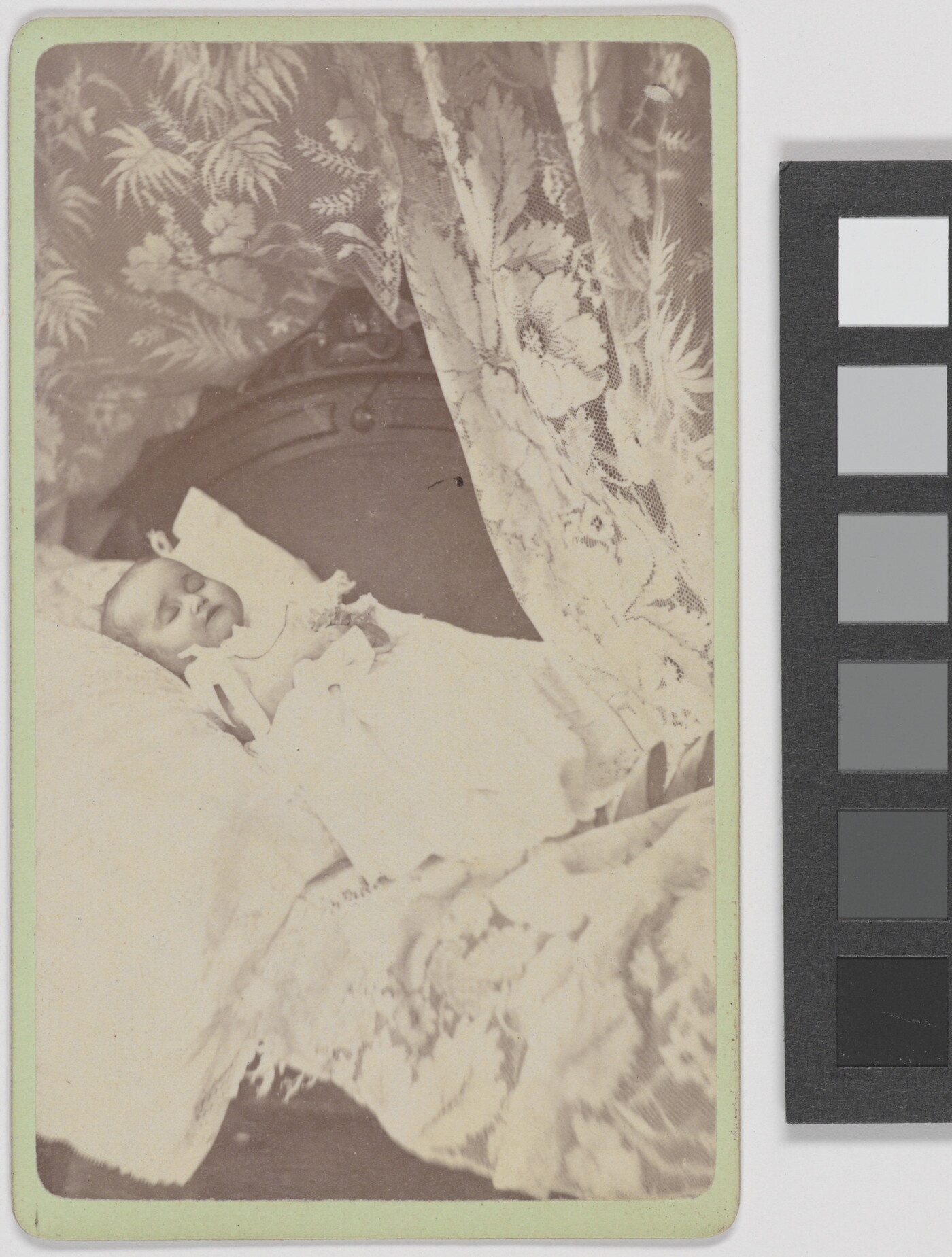 A black-and-white photograph of a White baby wearing a white gown and positioned on a pillow framed by a floral curtain.