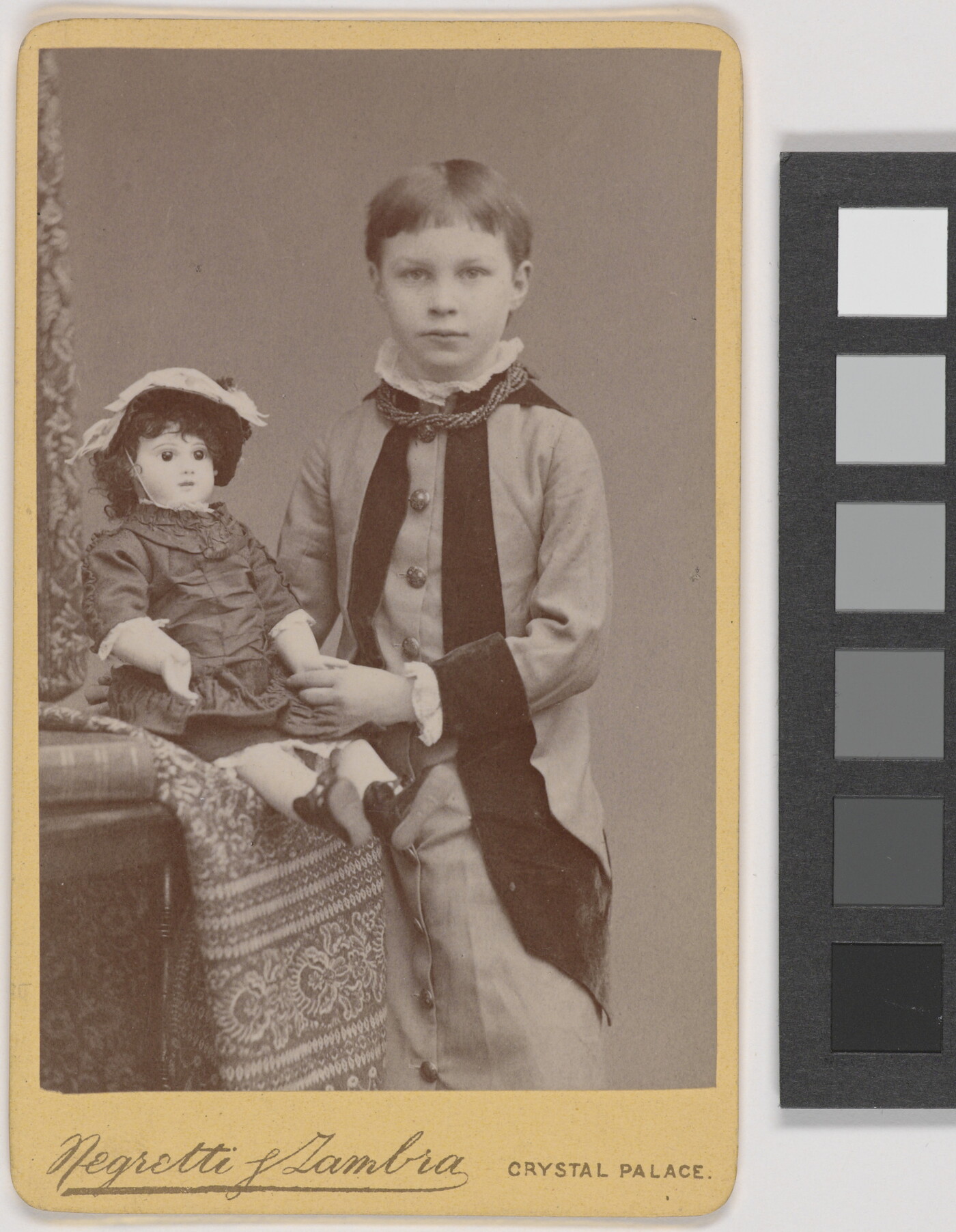 A black-and-white portrait of a young White child with short hair standing next to a seated doll.