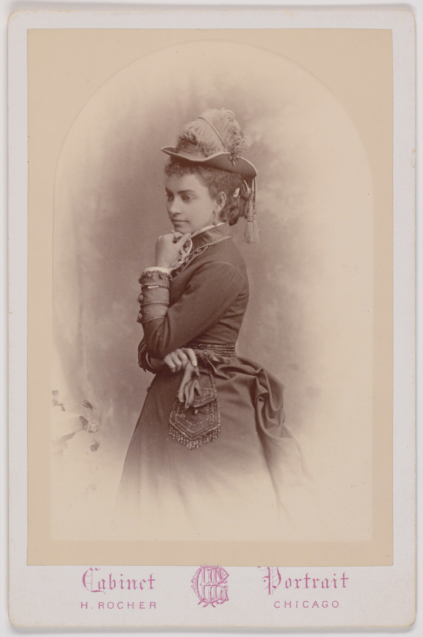 A black-and-white portrait photograph of a young White woman, standing in profile, wearing a hat decorated with feathers and tassels, holding a beaded purse in one hand, the other hand at her chin.