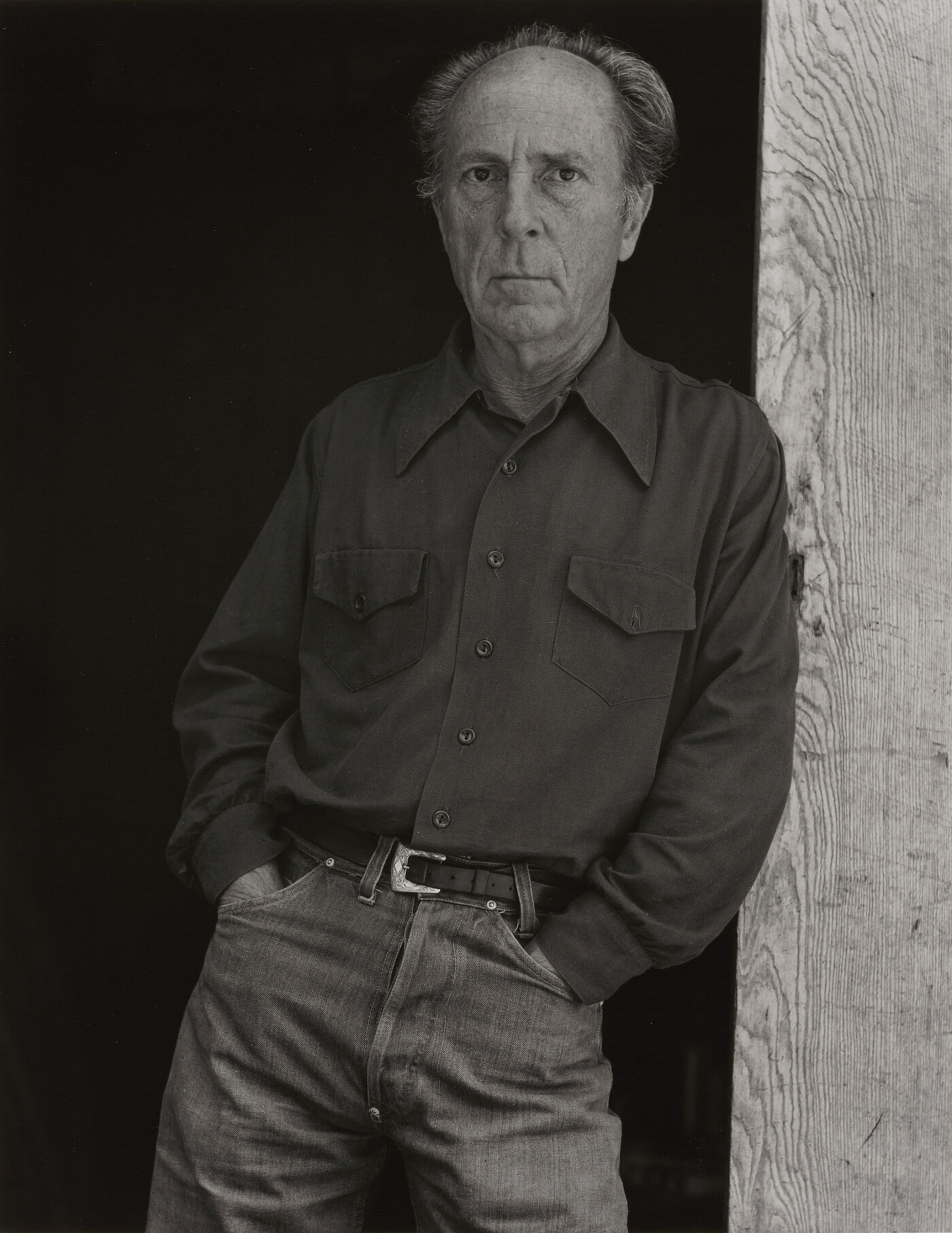 A black-and-white portrait of an older, balding White man wearing jeans and a long-sleeve button-down shirt leaning against a wooden beam.