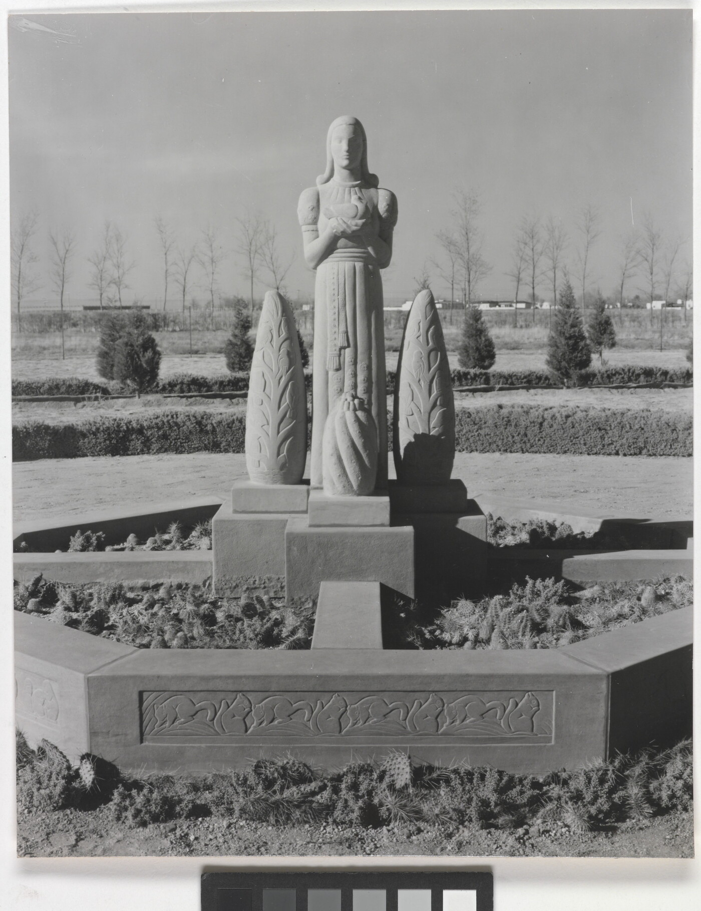 A black-and-white photograph of a stone statue of a stylized figure standing between stylized stone trees.