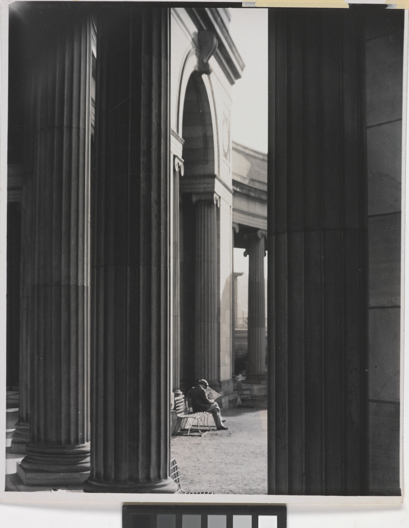 A black-and-white photograph of a person sitting on a bench reading a newspaper as seen from between large fluted columns.