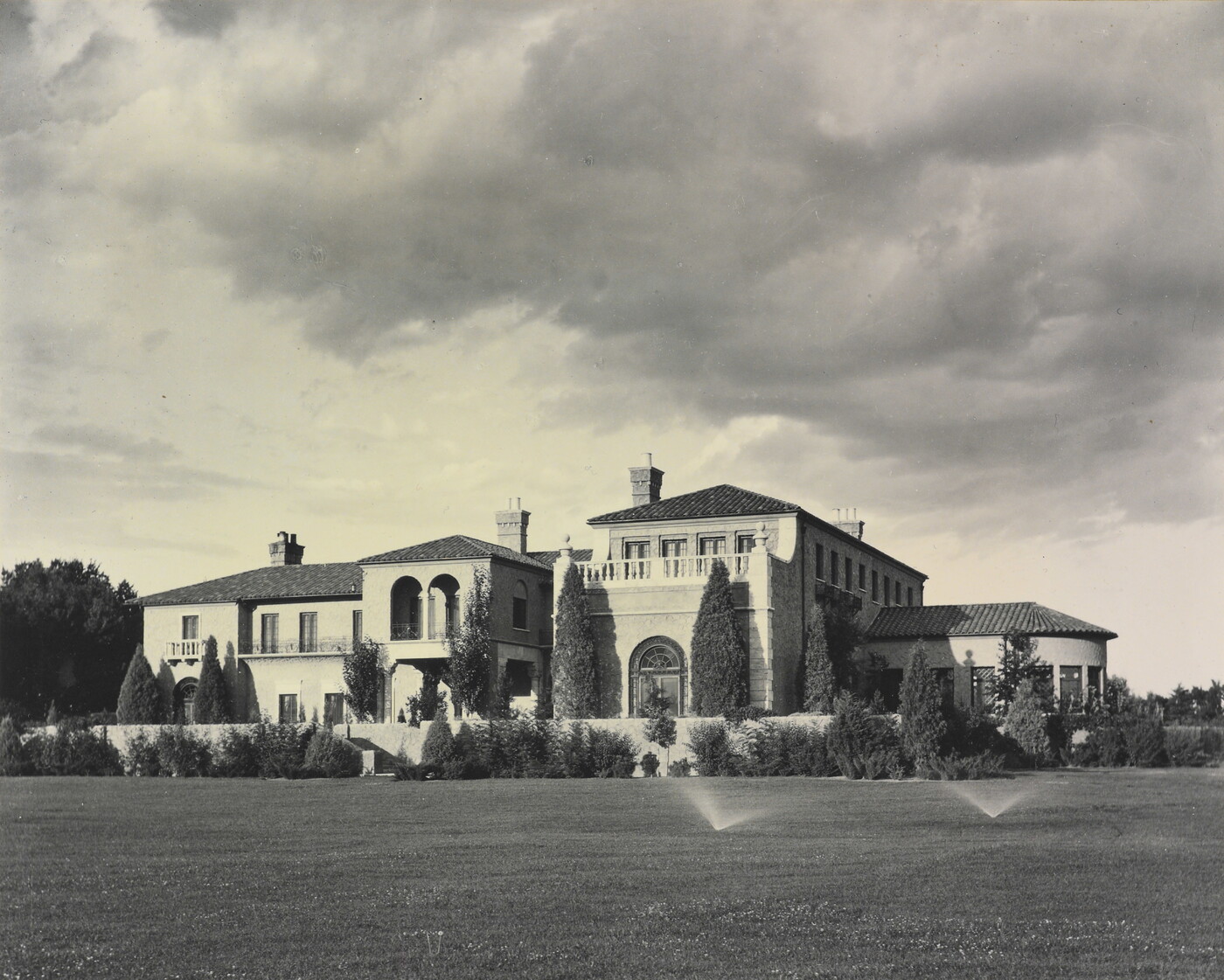 A black-and-white photograph of a large multi-story mansion with a mission-style roof and a large grass yard with sprinklers on.
