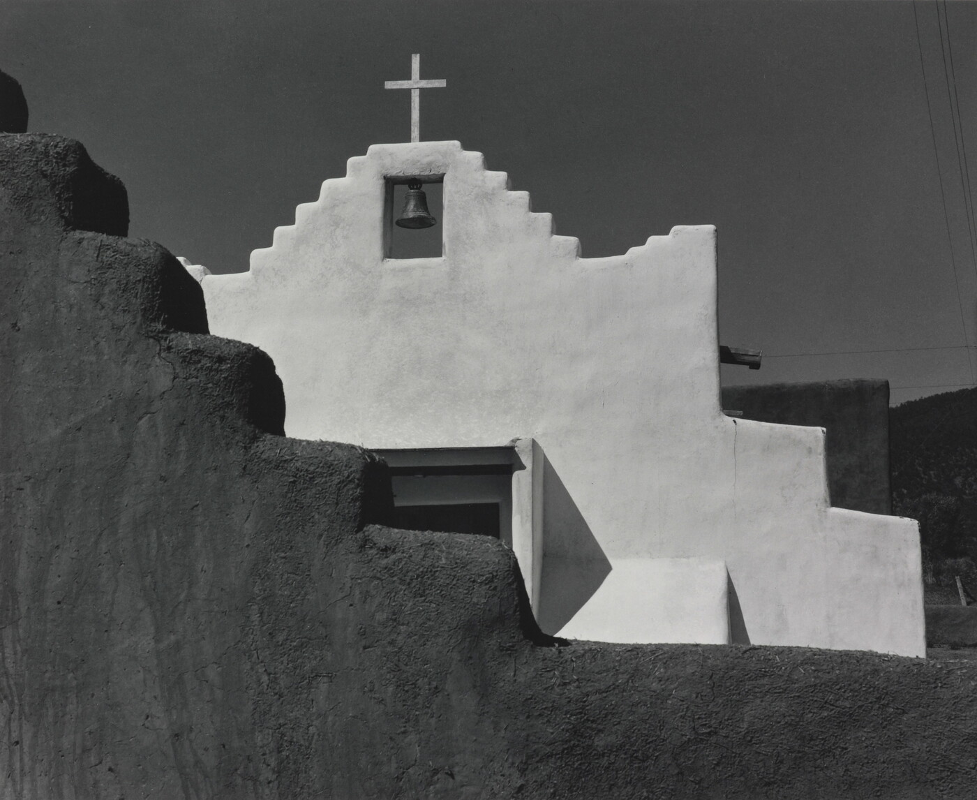A black-and-white photograph of a terraced adobe bell tower with a cross on top, partially hidden by a terraced adobe wall.