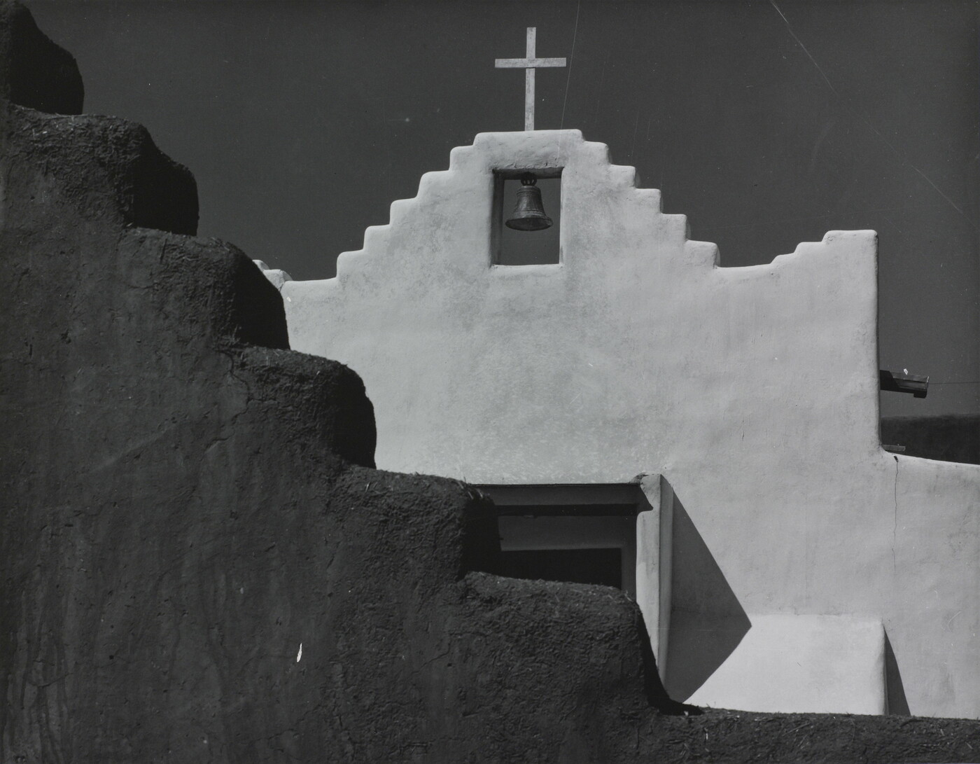 A black-and-white photograph of a terraced adobe bell tower with a cross on top, partially hidden by a terraced adobe wall.