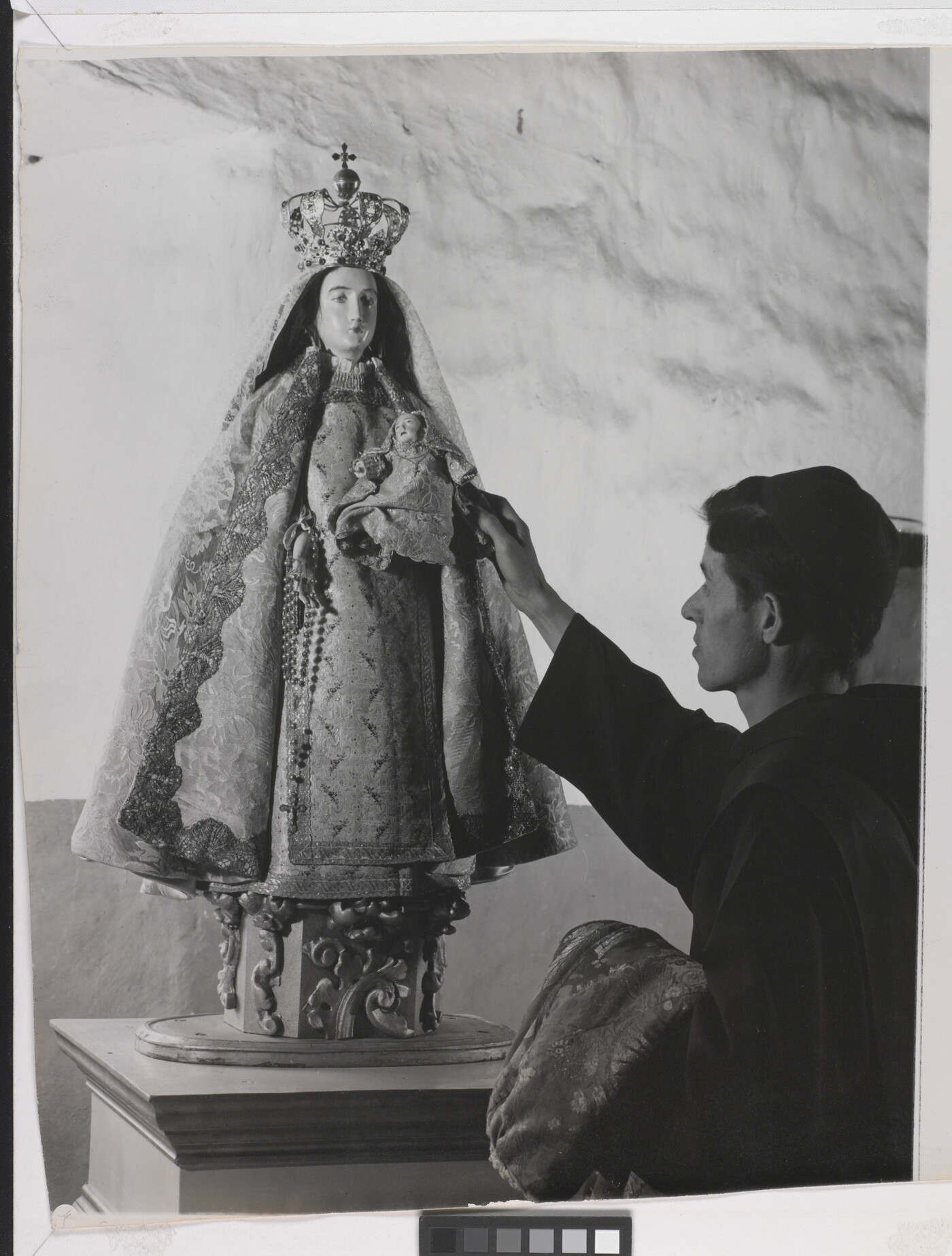 A black-and-white photograph of a friar touching a statue of the Virgin Mary dressed as queen of heaven holding the infant Jesus.