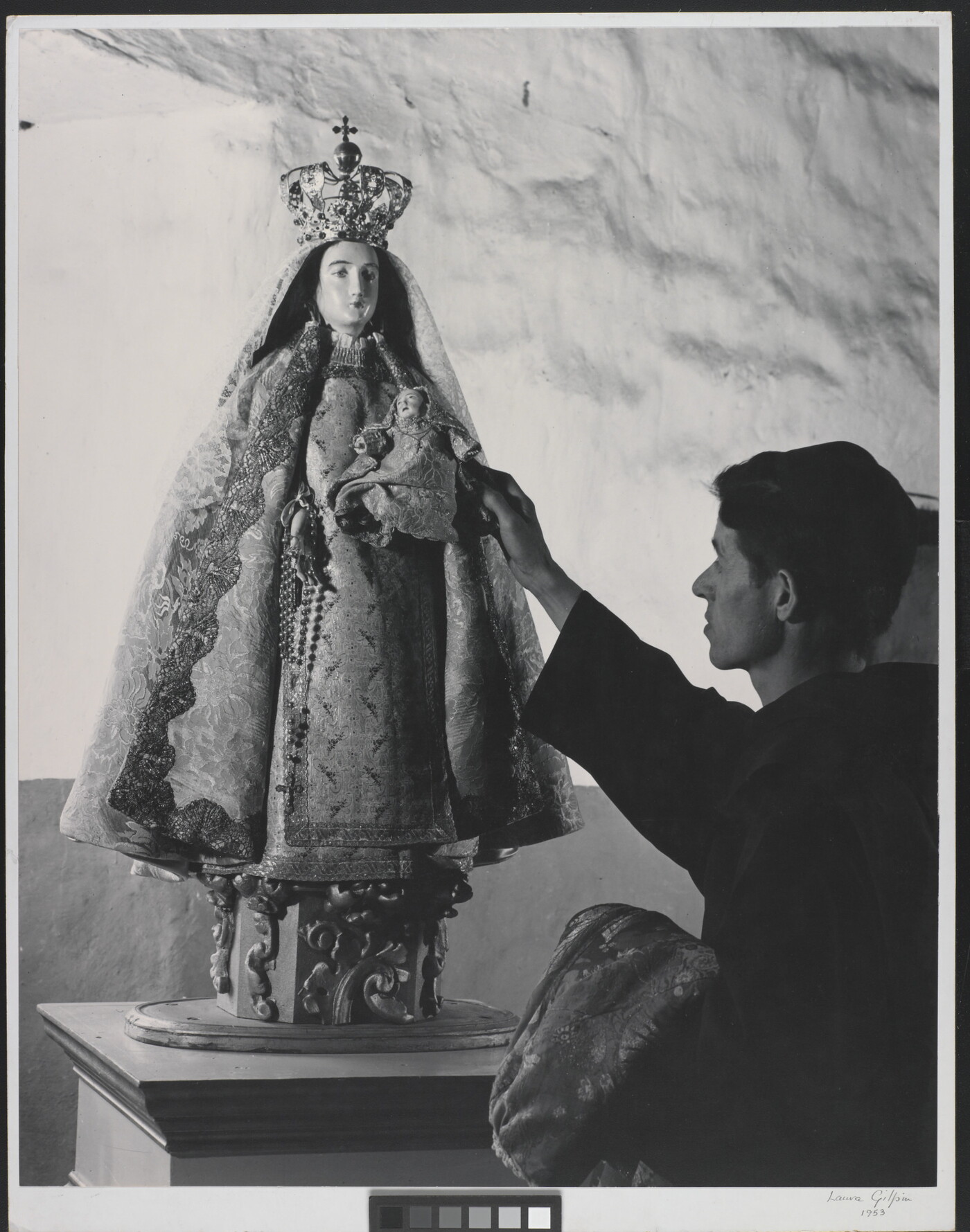 A black-and-white photograph of a friar touching a statue of the Virgin Mary dressed as queen of heaven holding the infant Jesus.