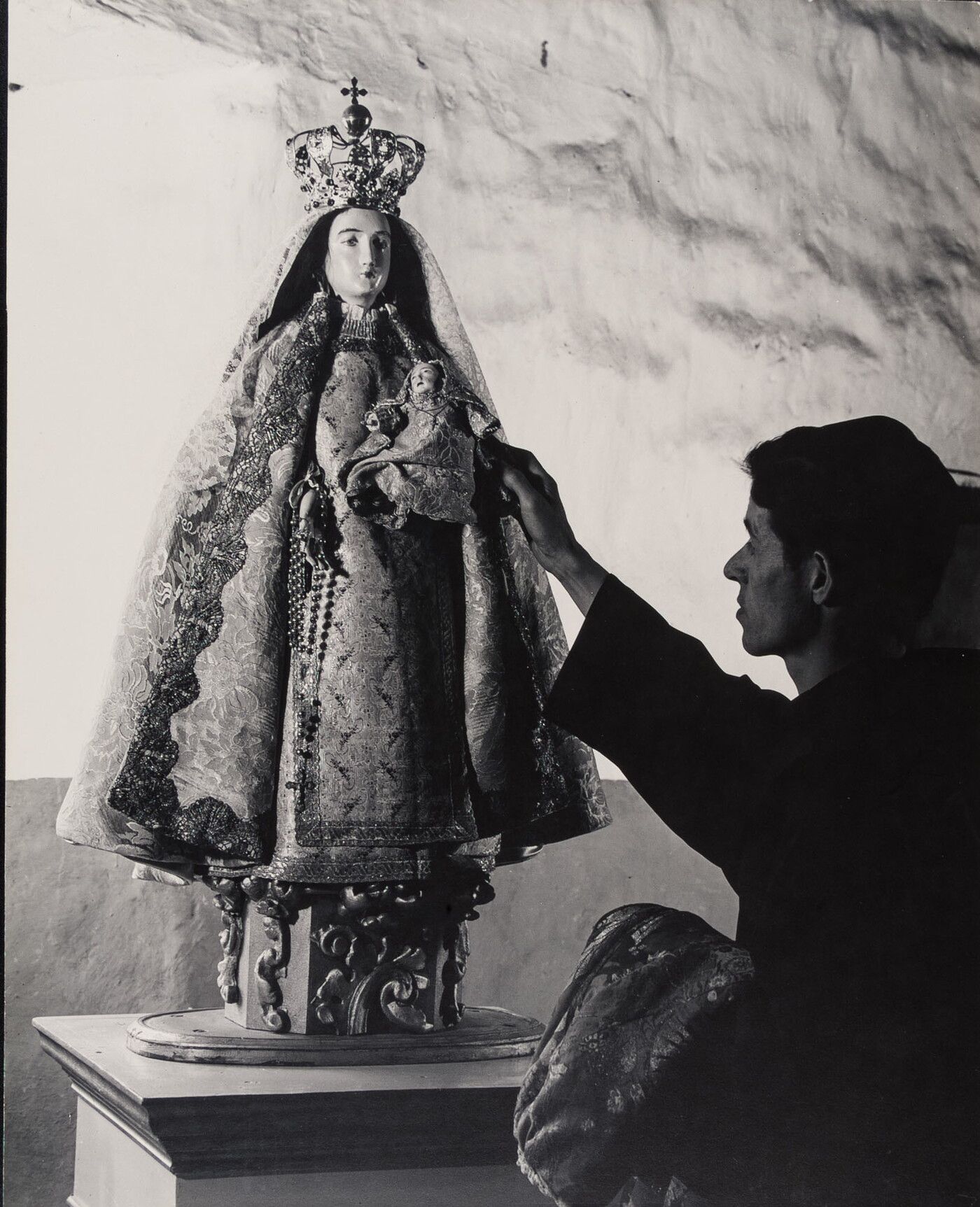 A black-and-white photograph of a friar touching a statue of the Virgin Mary dressed as queen of heaven holding the infant Jesus.