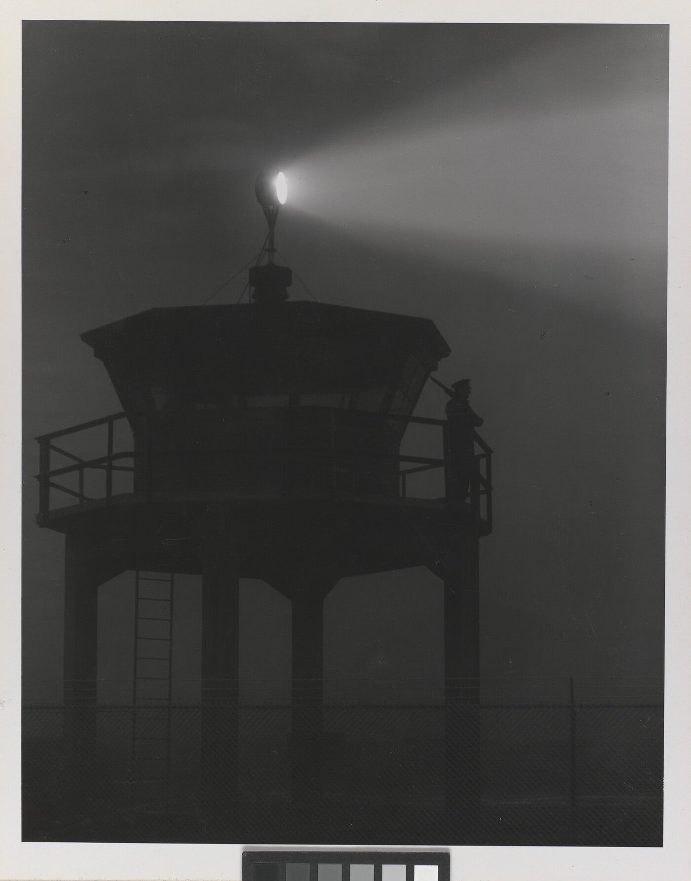 A black-and-white photograph of a person standing in silhouette atop a tower with a bright spotlight shining from the top.