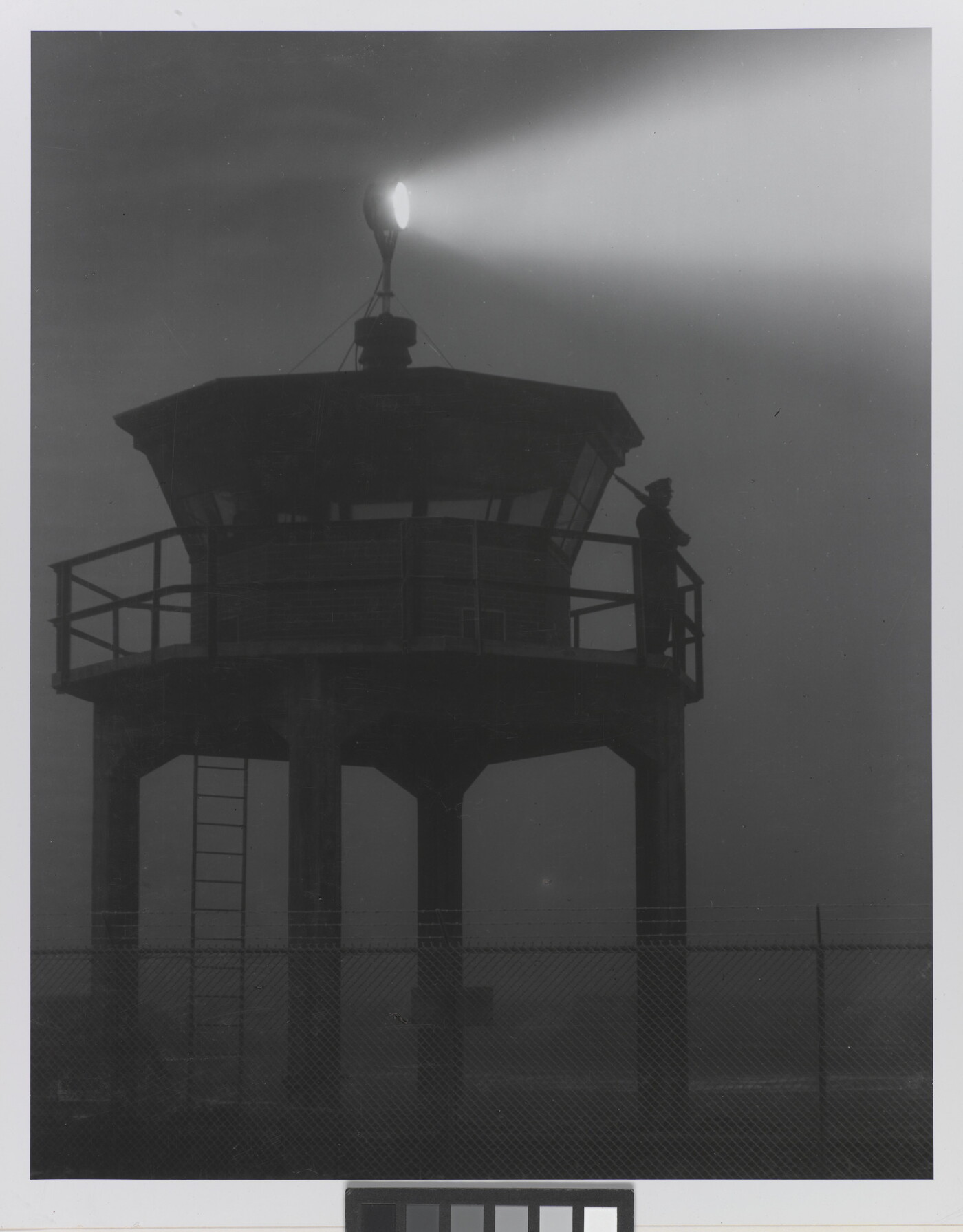 A black-and-white photograph of a person standing in silhouette atop a tower with a bright spotlight shining from the top.