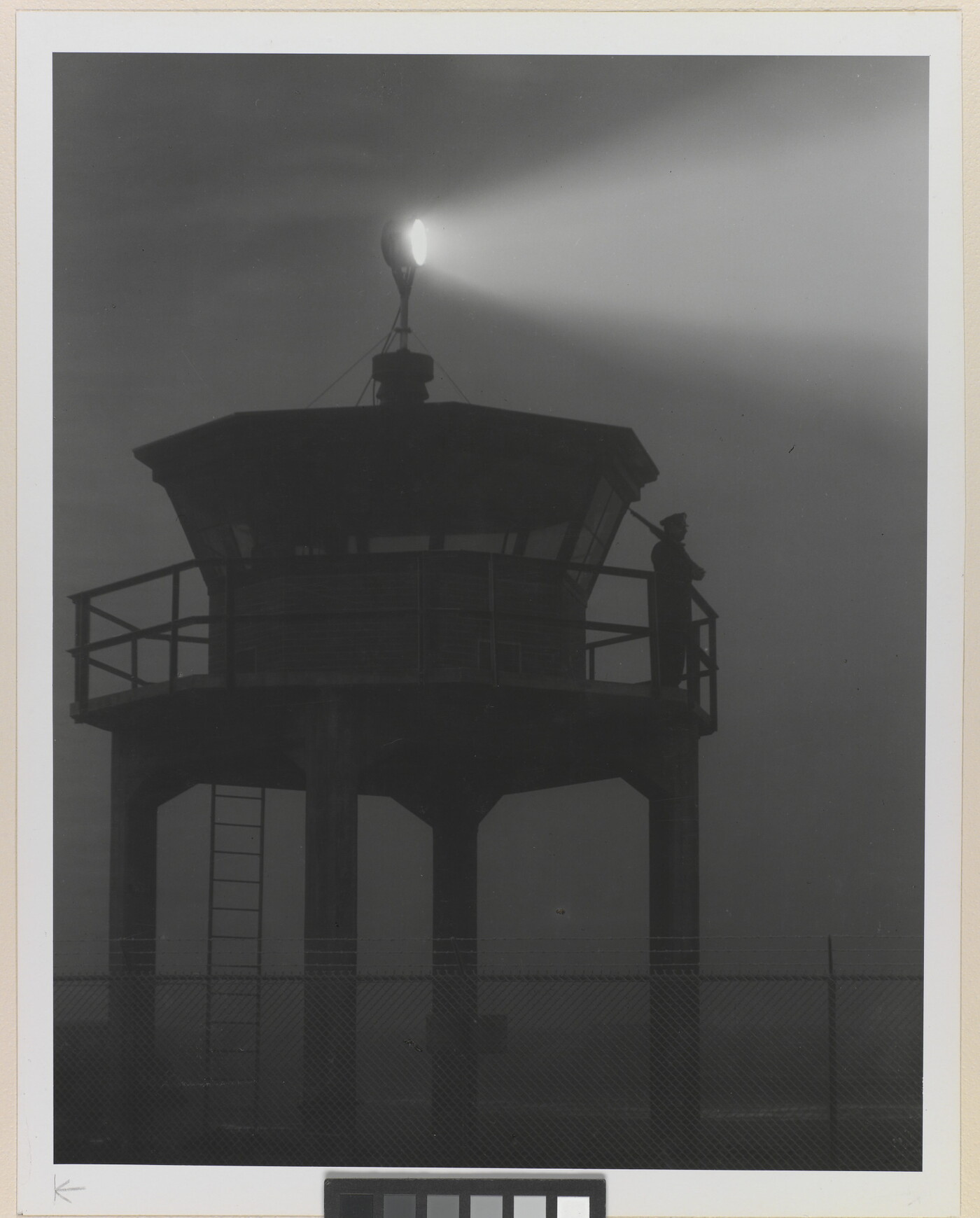 A black-and-white photograph of a person standing in silhouette atop a tower with a bright spotlight shining from the top.