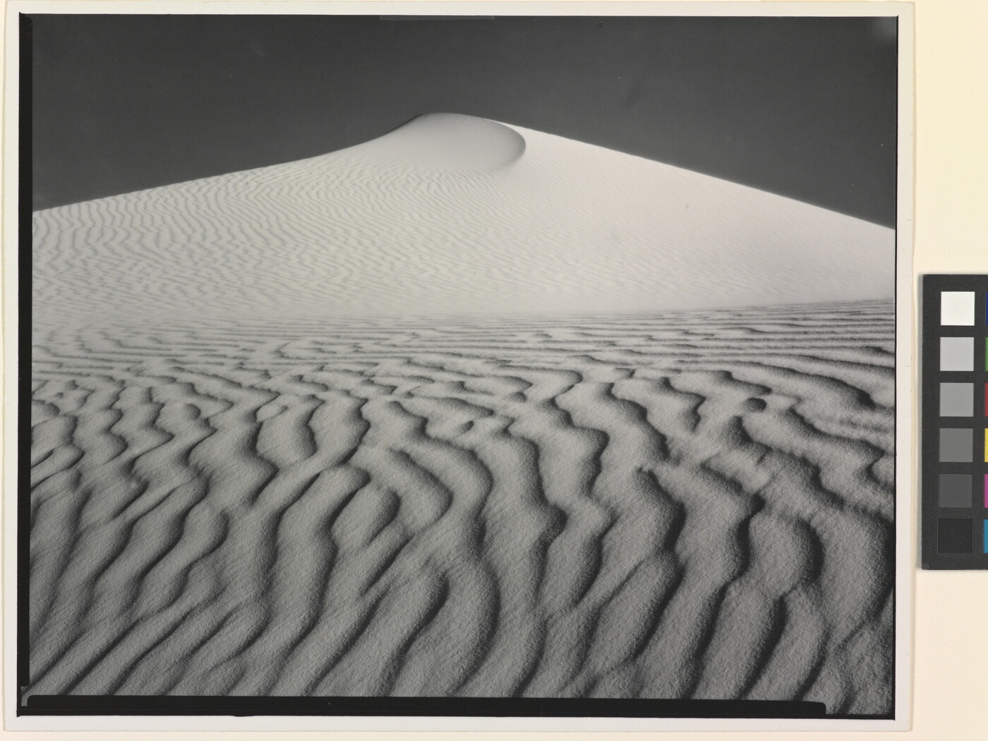 A sepia-toned photograph of a deeply-rippled sand dune rising to a point into the sky.
