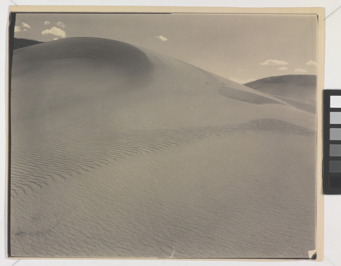 A sepia-toned photograph of a rippling sand dune rising up into a sky dotted with a few small clouds.