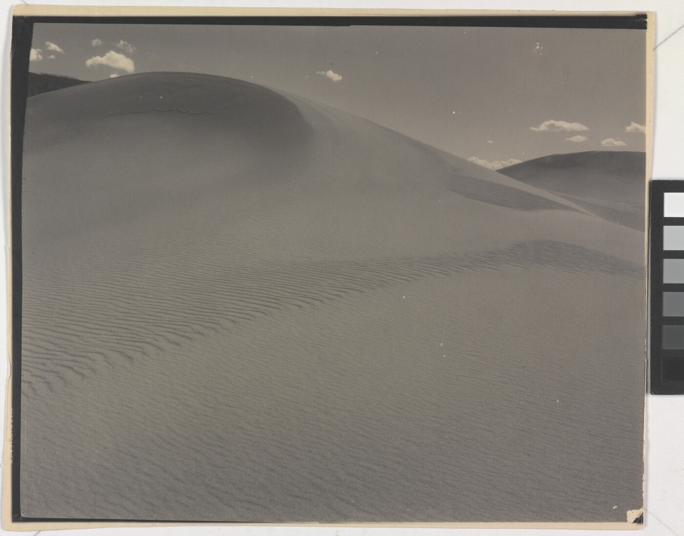 A sepia-toned photograph of a rippling sand dune rising up into a sky dotted with a few small clouds.