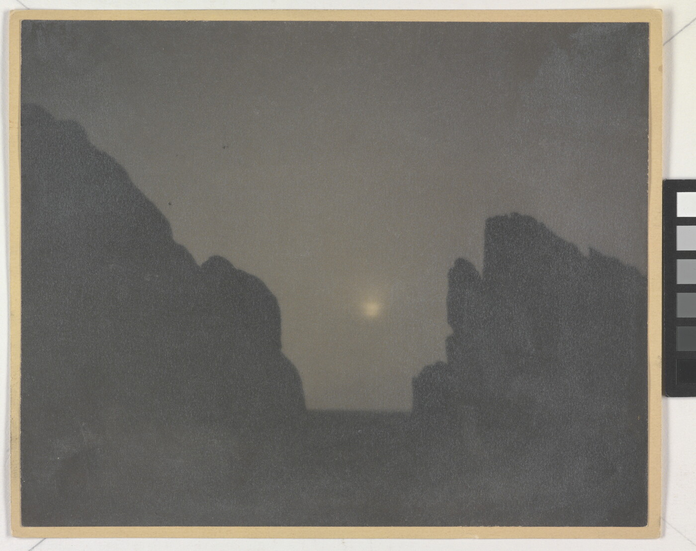 A black-and-white photograph of two rock formations in silhouette with a hazy moon in between them.