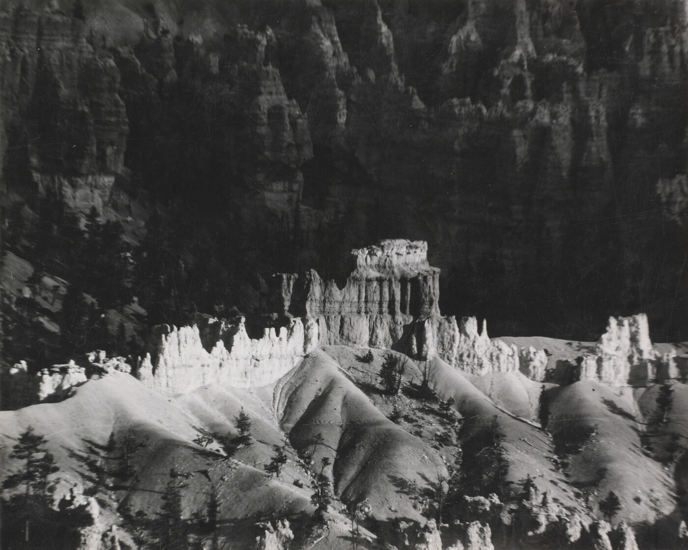 A black-and-white photograph of a landscape with hoodoos, or vertical spires of rock, on top of sand dune shapes with cliffs in the background.