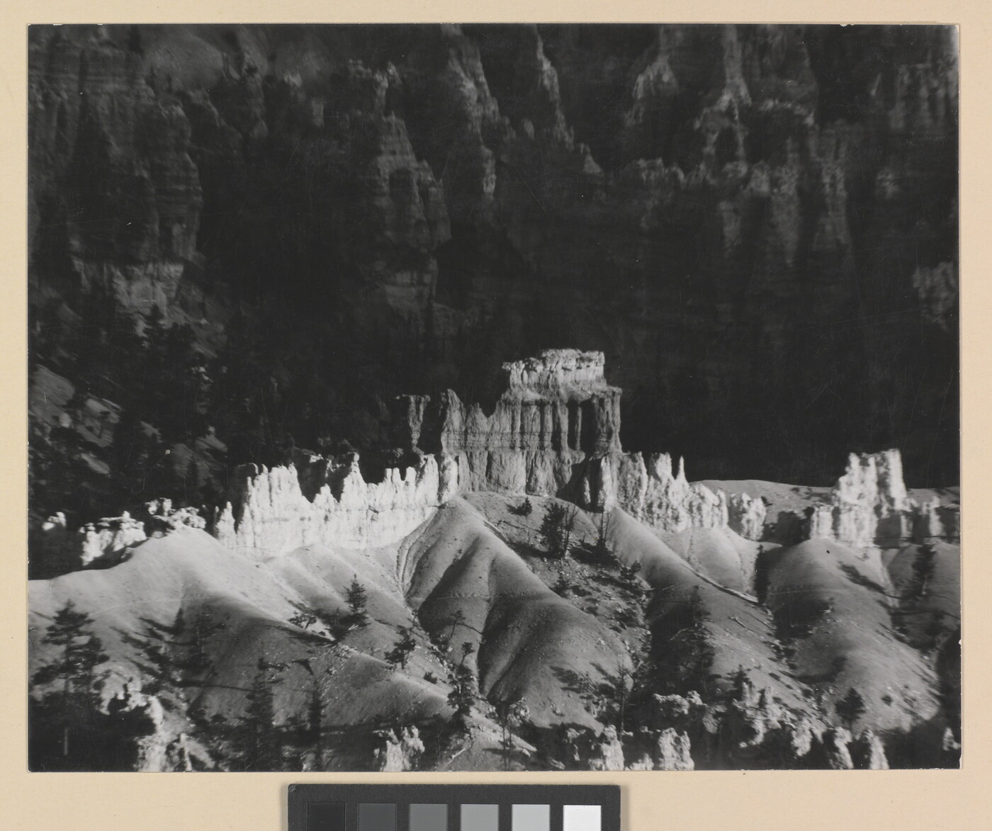 A black-and-white photograph of a landscape with hoodoos, or vertical spires of rock, on top of sand dune shapes with cliffs in the background.