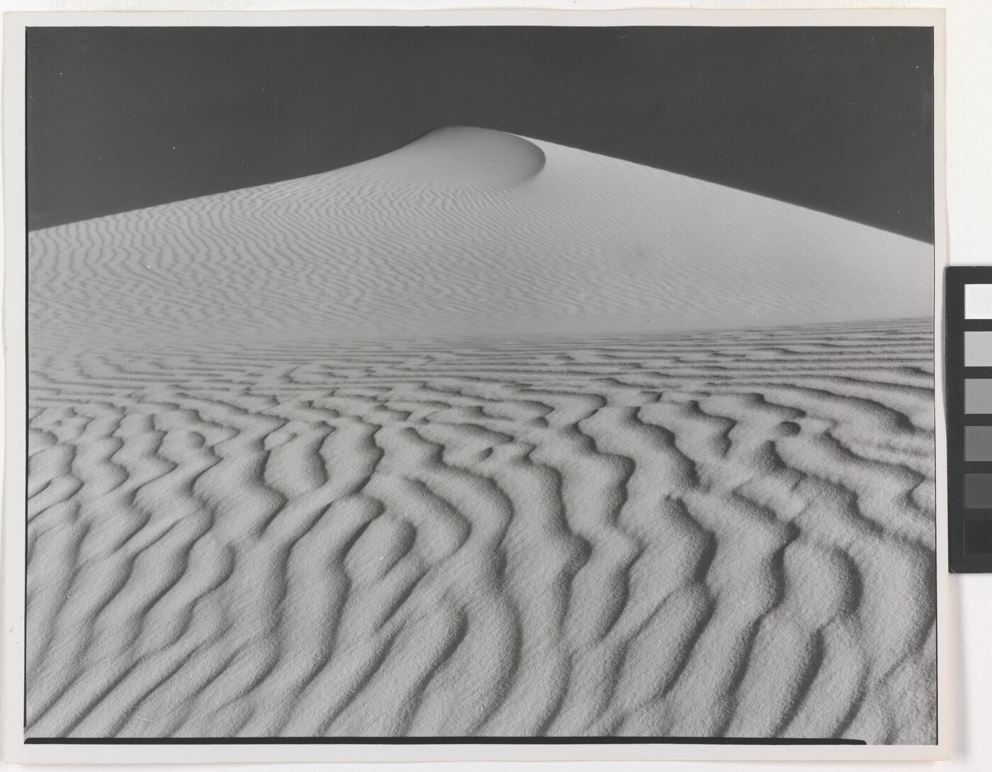 A sepia-toned photograph of a deeply-rippled sand dune rising to a point into the sky.