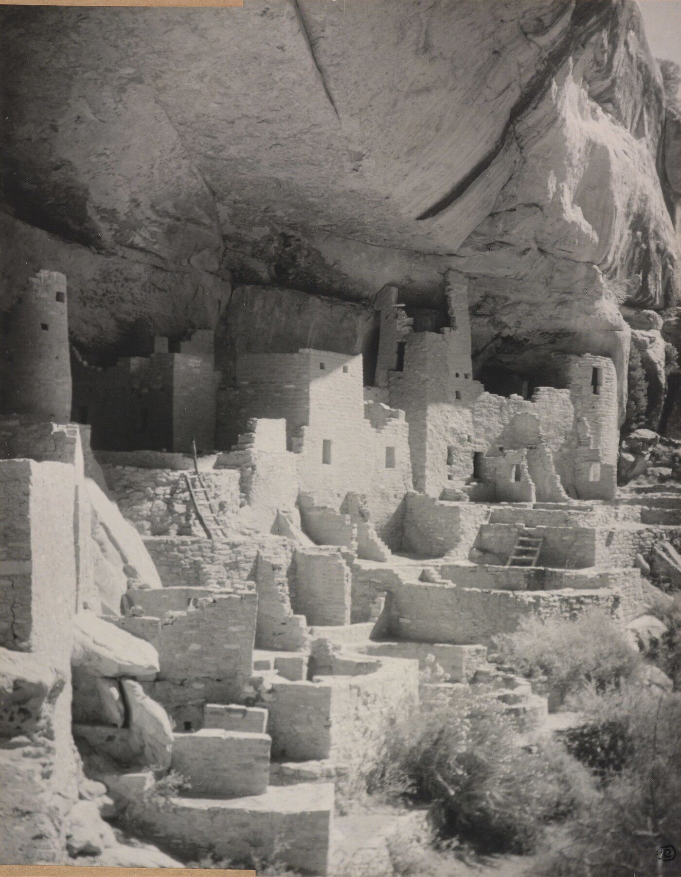 A black-and-white photograph of an abandoned, crumbling rock city built into the side of a desert cliff.