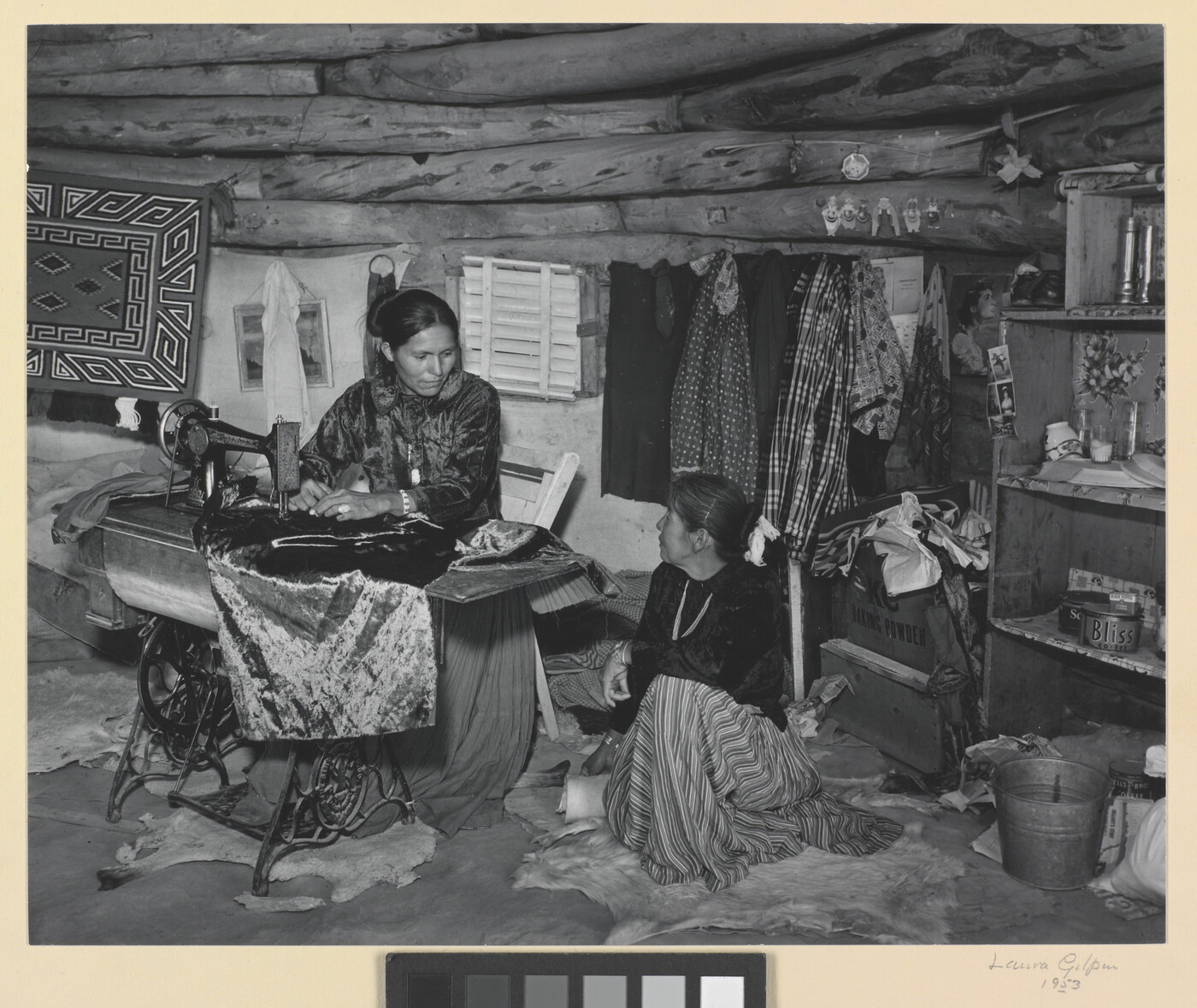 A black-and-white photograph of two Indigenous women seated, talking, and sewing inside of a low-ceilinged, wood structure.