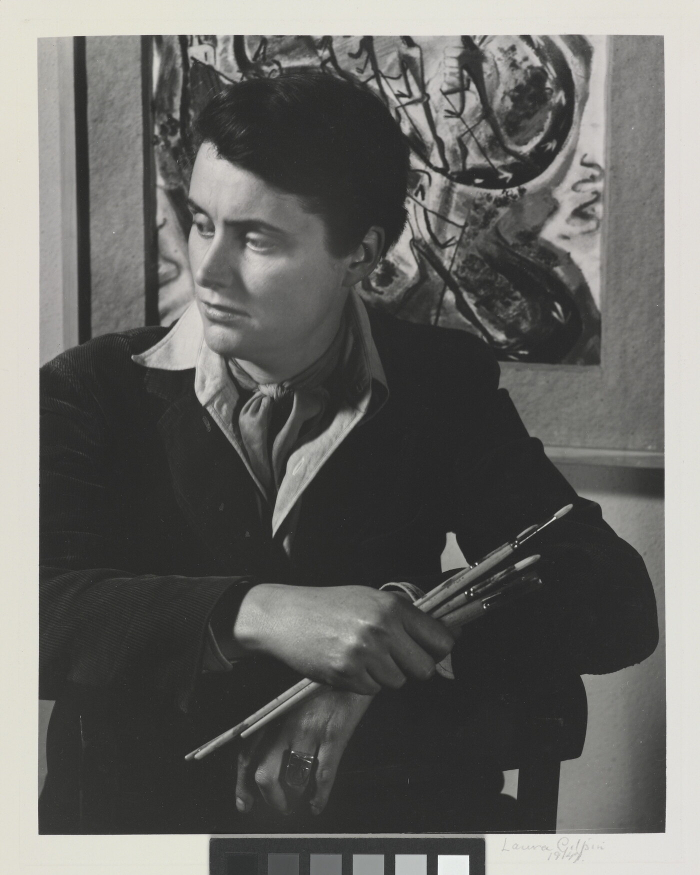 A black-and-white portrait photograph of a seated White woman holding paintbrushes in front of an abstract painting.