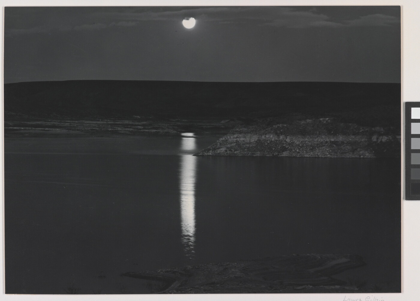 A black-and-white photograph of a night sky and full moon reflected across calm water.