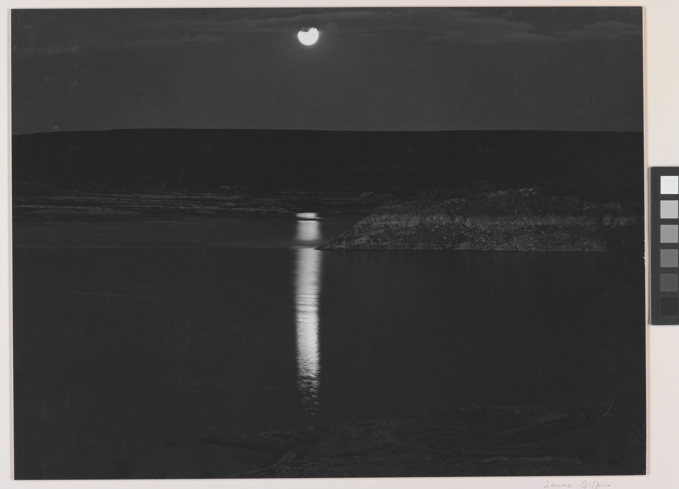 A black-and-white photograph of a night sky and full moon reflected across calm water.