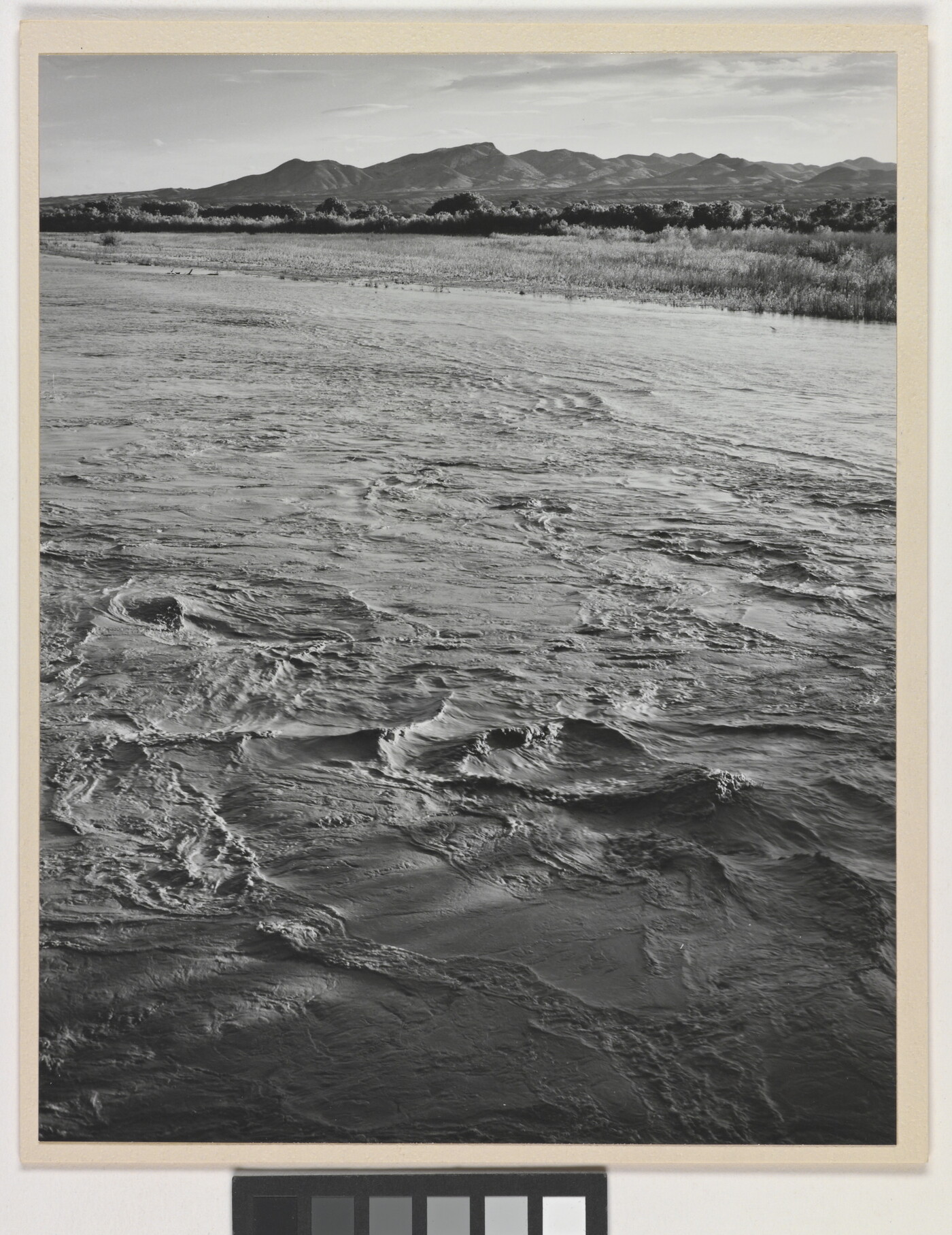 A black-and-white photograph of a choppy river and then grassy plains and mountains beyond the shore.