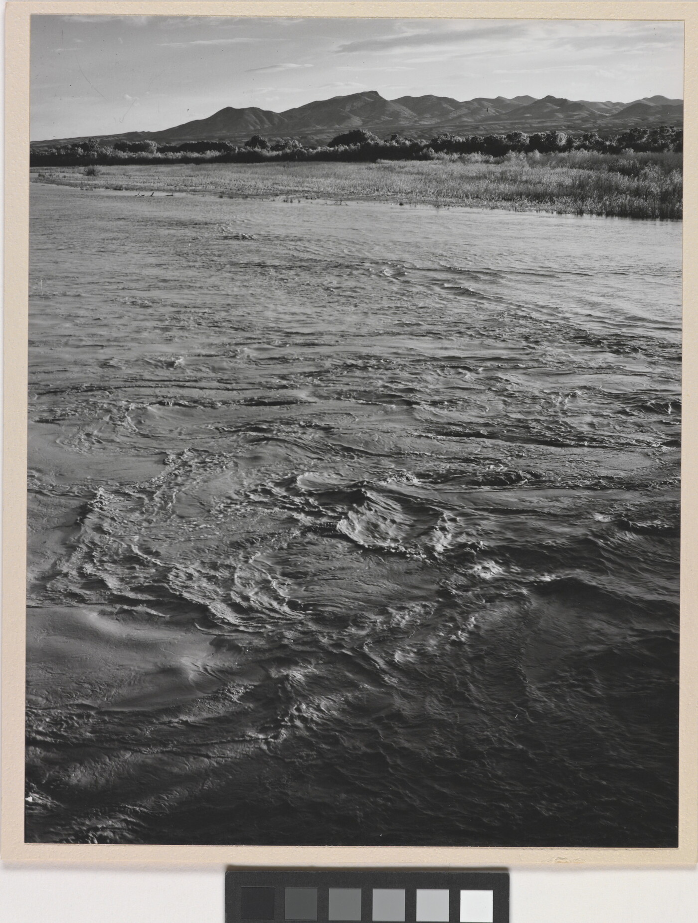 A black-and-white photograph of a choppy river and then grassy plains and mountains beyond the shore.