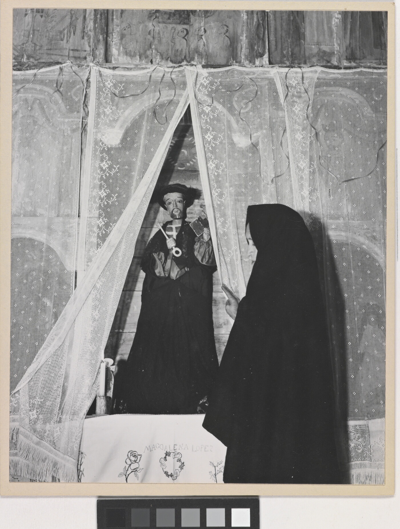 A black-and-white photograph of a woman wearing a black shawl over her head facing a statue of a saint holding a large key.