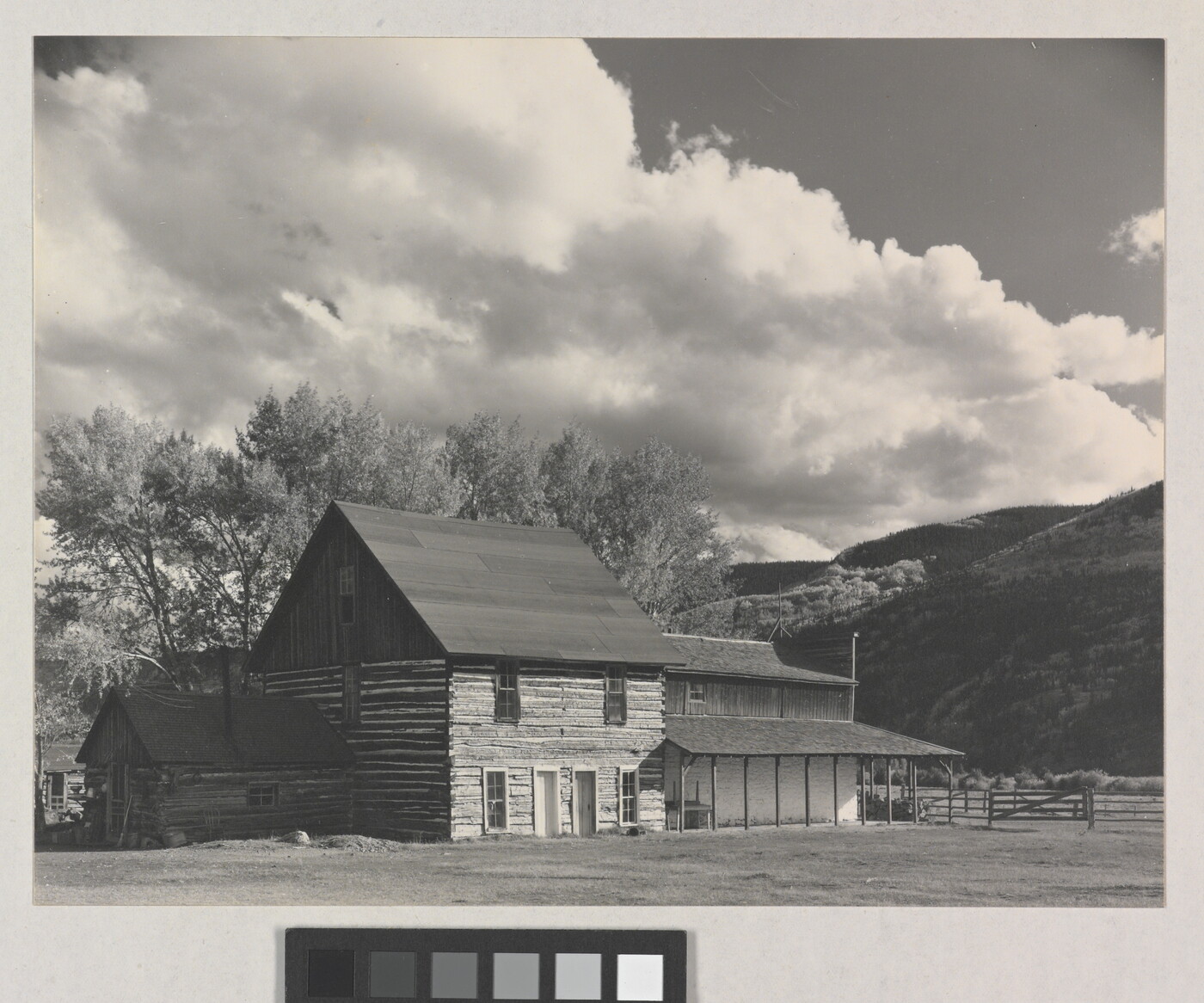 A black-and-white photograph of a three-story log building with mountains and large puffy clouds in the background.