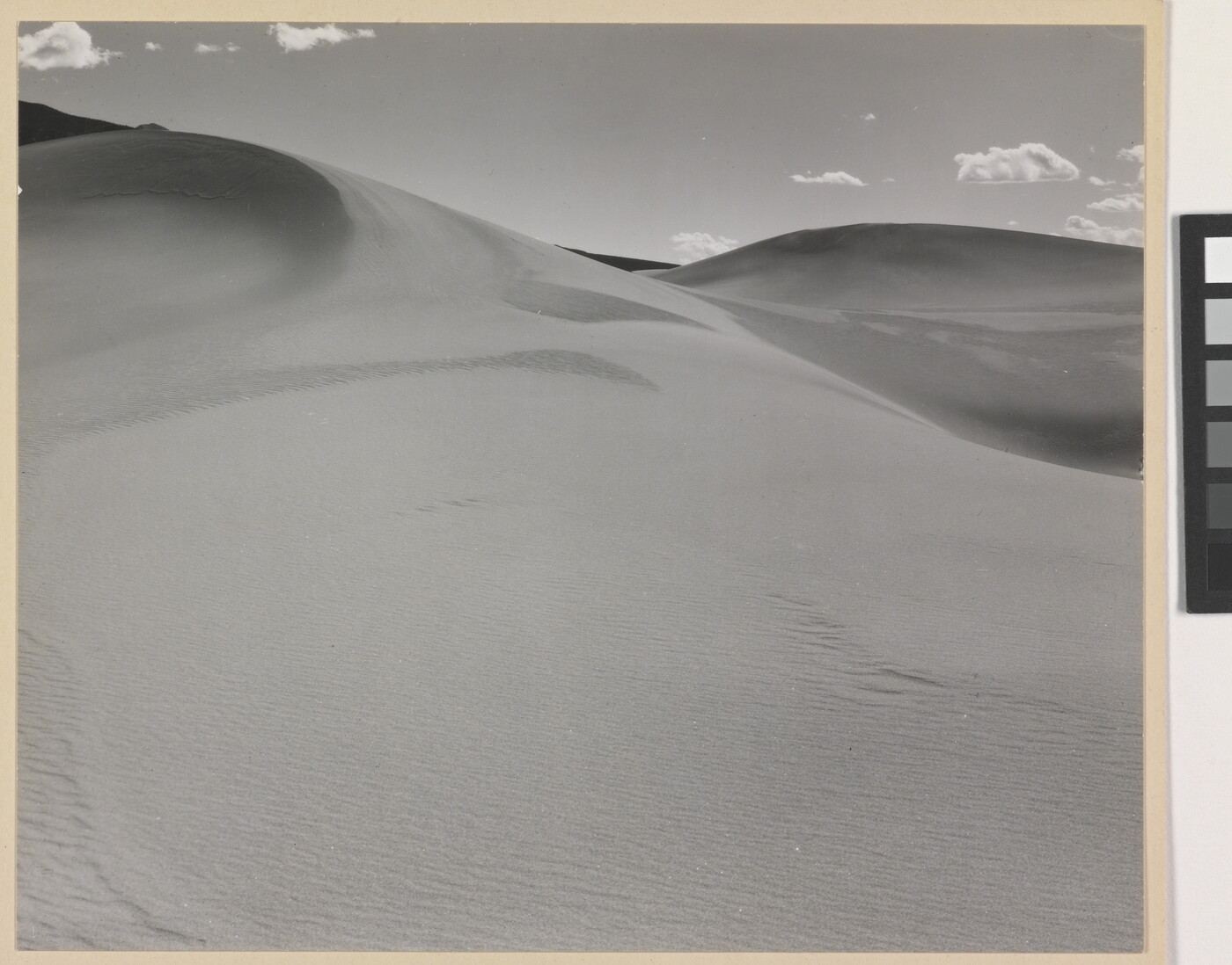 A black-and-white photograph of a rippling sand dune rising up into a sky dotted with a few small clouds.