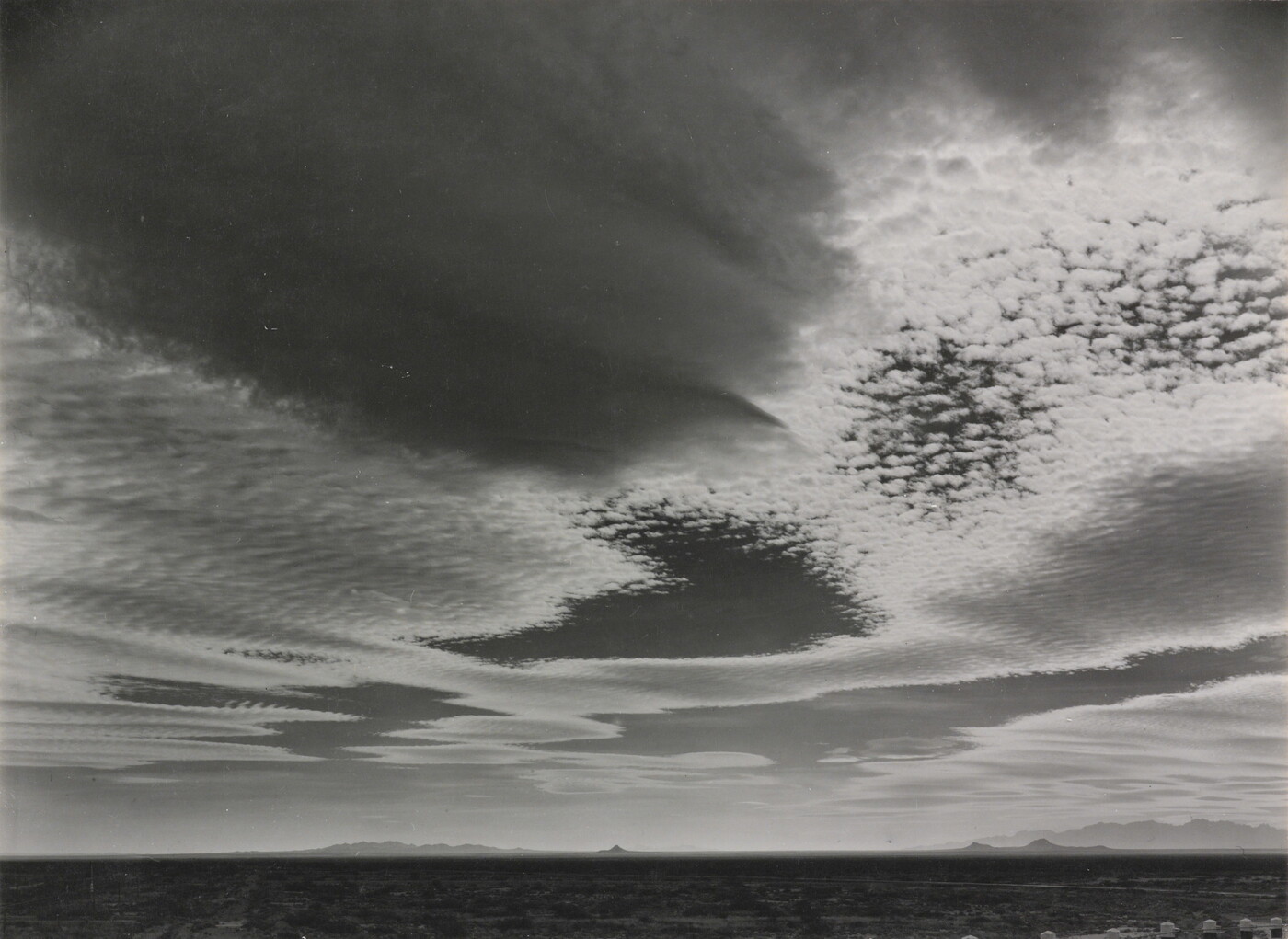 A black-and-white photograph of an expansive desert plain under rippled clouds with mountains in the far distance.