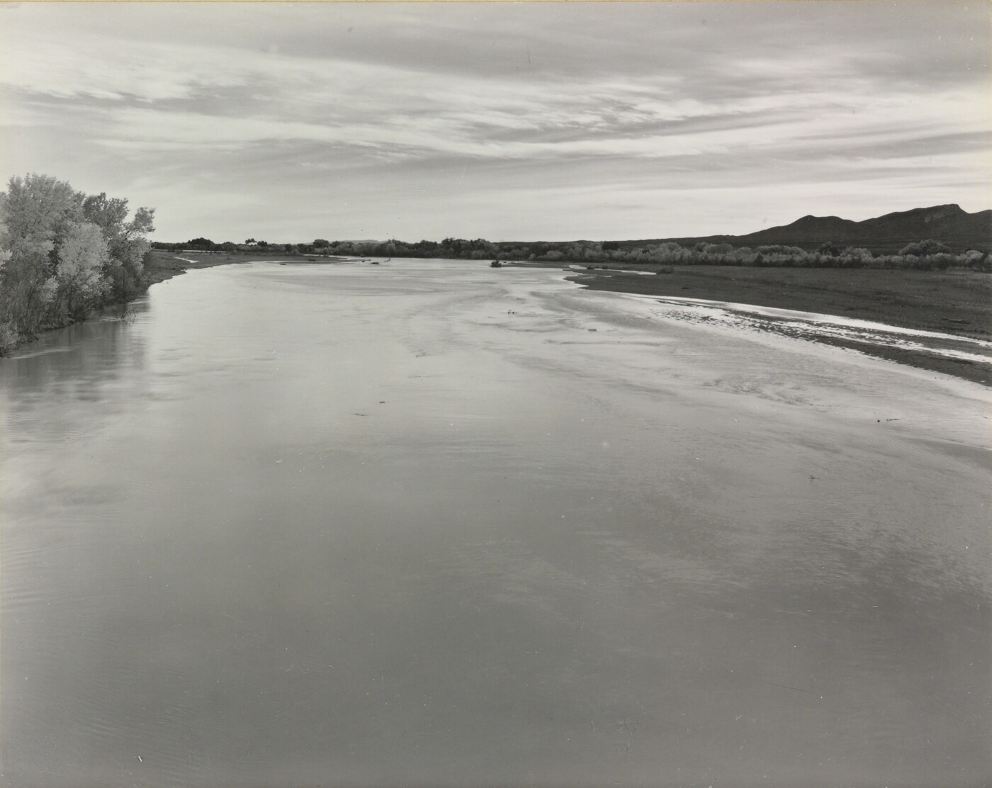 A black-and-white photograph of a calm river with trees on the left shore and mountains in the distance.