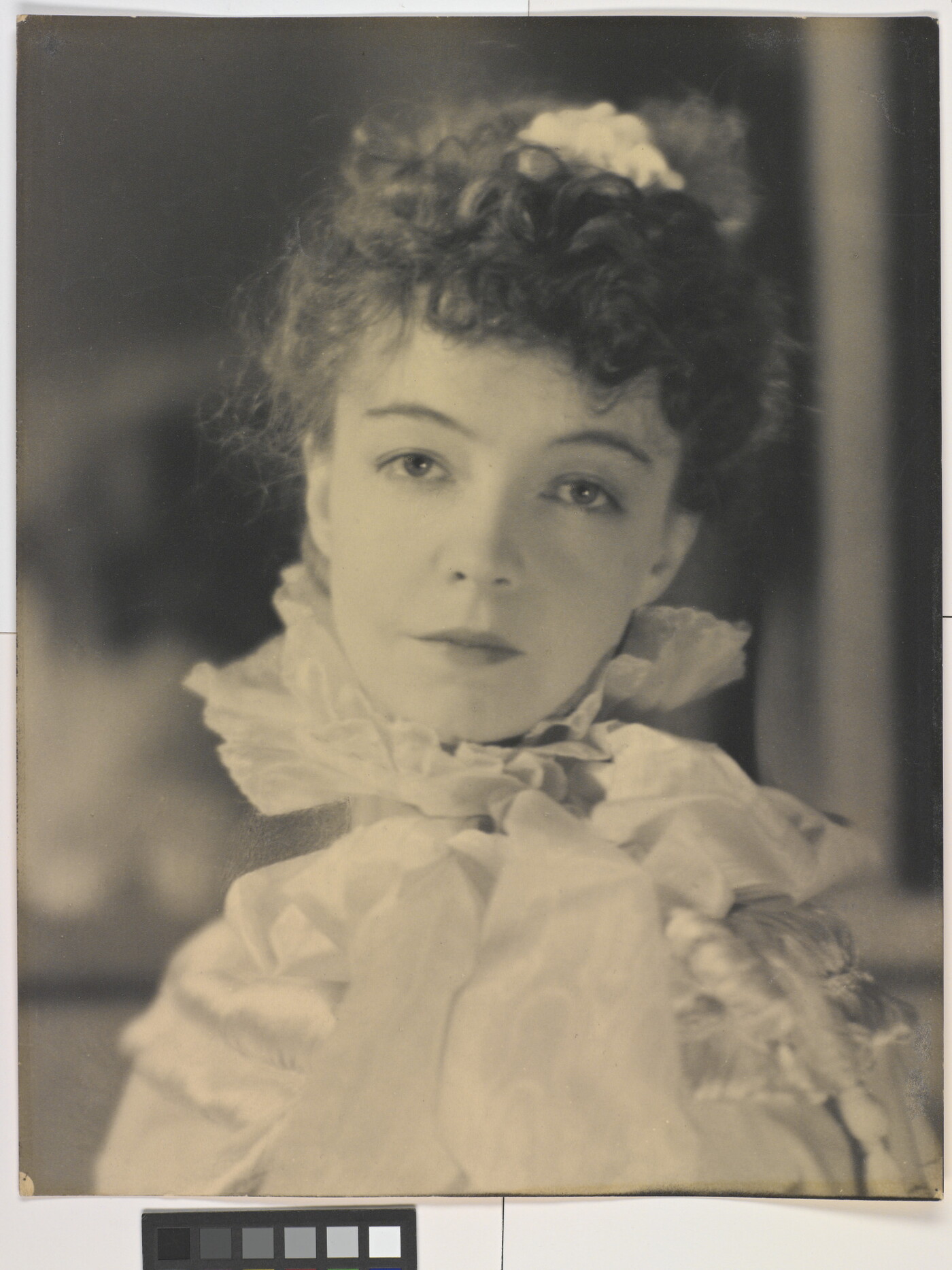 A black-and-white portrait photograph of a White woman with curly hair pulled on top of her head, wearing a top with a ruffled collar.