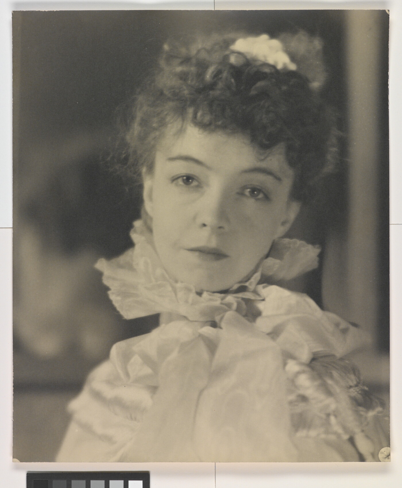 A black-and-white portrait photograph of a White woman with curly hair pulled on top of her head, wearing a top with a ruffled collar.