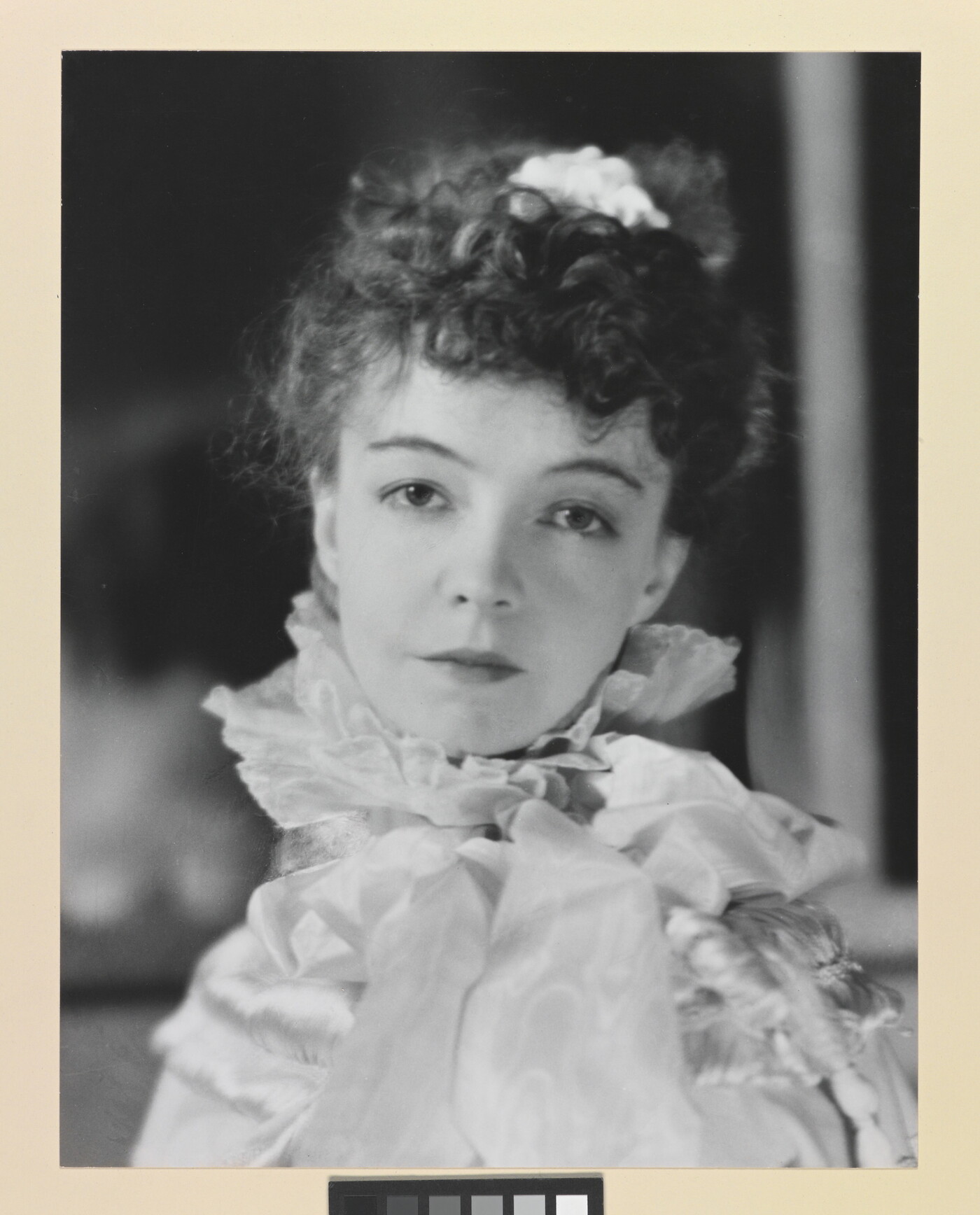 A black-and-white portrait photograph of a White woman with curly hair pulled on top of her head, wearing a top with a ruffled collar.